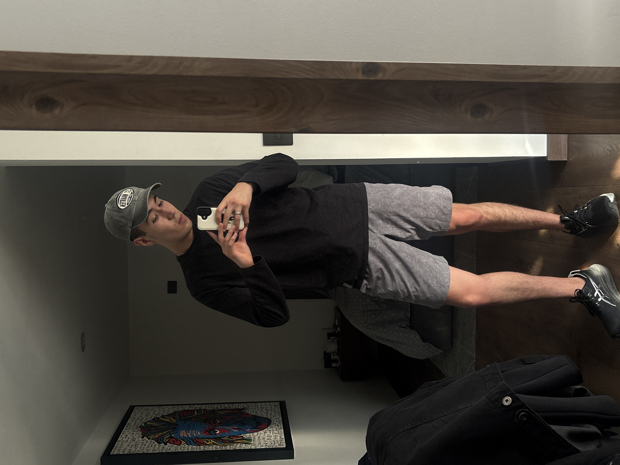 The image features a young man captured in a mirror selfie within an indoor setting. He stands upright, holding a light-colored smartphone with his left hand to take the photo. He is wearing a grey baseball cap with a circular logo, a dark long-sleeved shirt, and light grey patterned shorts. A ring is visible on his left hand. His feet are clad in black athletic shoes with white accents.
The background, seen through the mirror reflection, includes a dark wooden shelf or beam positioned above his head, with a lighter wall segment visible higher up. The floor is also dark wood. To his right, on the floor, rests a rectangular framed piece of artwork, which depicts a colorful, stylized face against a background of intricate script or pattern. A folded dark jacket or garment lies on the floor to his left.
The activity is a casual mirror self-portrait. The man's expression is neutral as he looks downwards, presumably at his phone screen. Notable details include the mirrored text on his baseball cap's logo, which appears as "32 NAYLTED" but would likely be "DETRAAN 32" when un-mirrored. The indoor lighting is subdued, typical of an interior space. The overall scene suggests a contemporary, private room, possibly in San Bernardino Tlaxcalancingo, Mexico.