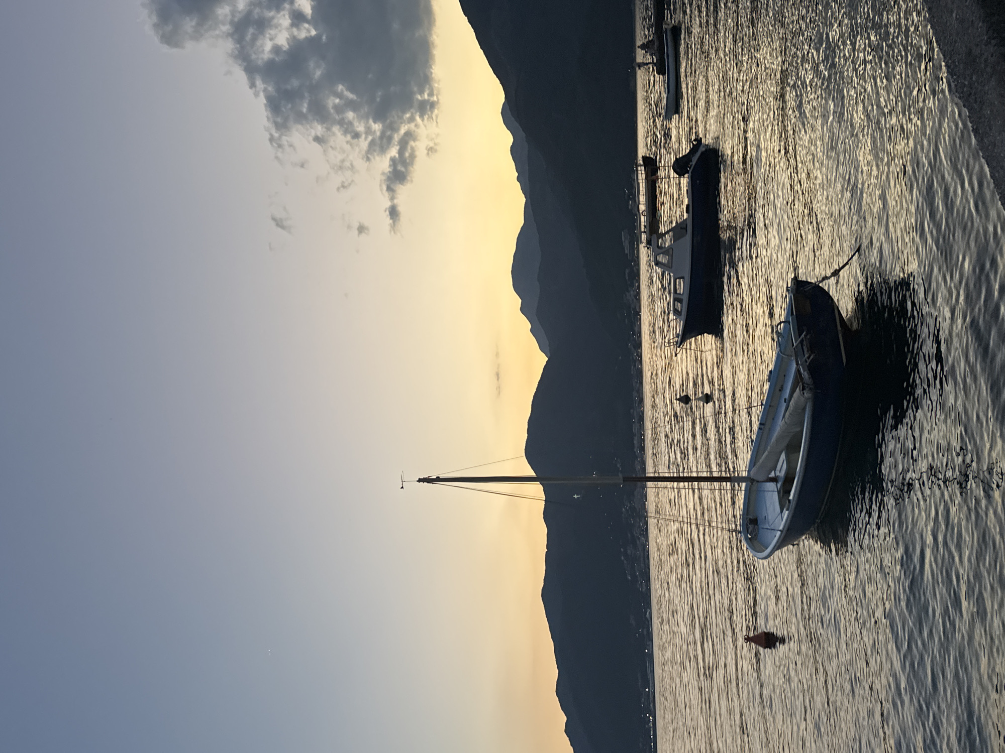 The image depicts a serene coastal scene at dusk. In the foreground, a small blue sailboat is moored in calm water, its mast reaching towards the sky. A red buoy floats nearby. In the middle ground, several other boats, including a motorboat, are anchored on the water. The background is dominated by the silhouetted outline of dark, undulating mountains against a gradient sky transitioning from a soft yellow at the horizon to a pale blue overhead. Scattered clouds add texture to the upper portion of the sky. The overall impression is one of tranquility and natural beauty.