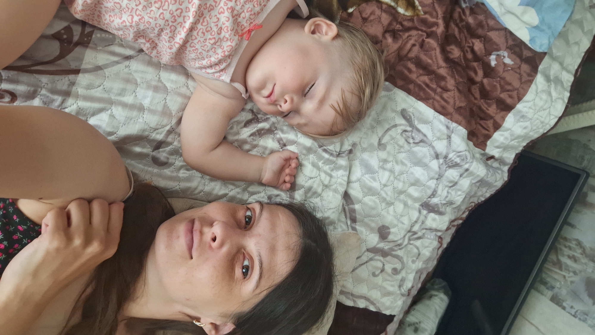 An adult female and a sleeping toddler are depicted lying on a quilted bedspread in an indoor setting, reportedly in Надеждино, Russia. The adult female, positioned on the right side of the frame, possesses dark brown hair and brown eyes, maintaining direct eye contact with the camera. She wears a light-colored hoop earring in her visible right ear and a dark-colored top featuring a floral pattern. Her right arm is partially visible, with her hand positioned near her shoulder. To her left, a toddler with short, light blonde hair is asleep, lying on their back with eyes closed and mouth slightly agape. The child wears a light-colored sleeveless top adorned with a pattern of small pink hearts and a small pink bow on the left shoulder strap. The toddler's left arm is bent at the elbow, with the hand resting on the bedspread. The bedspread itself is quilted, displaying sections of light gray with a dark floral or abstract pattern alongside sections of solid brown. In the upper right background, a dark, rectangular object, consistent with a television or monitor screen, is partially visible, accompanied by a light-colored, rolled object. The scene conveys a quiet moment of rest within a bedroom environment.