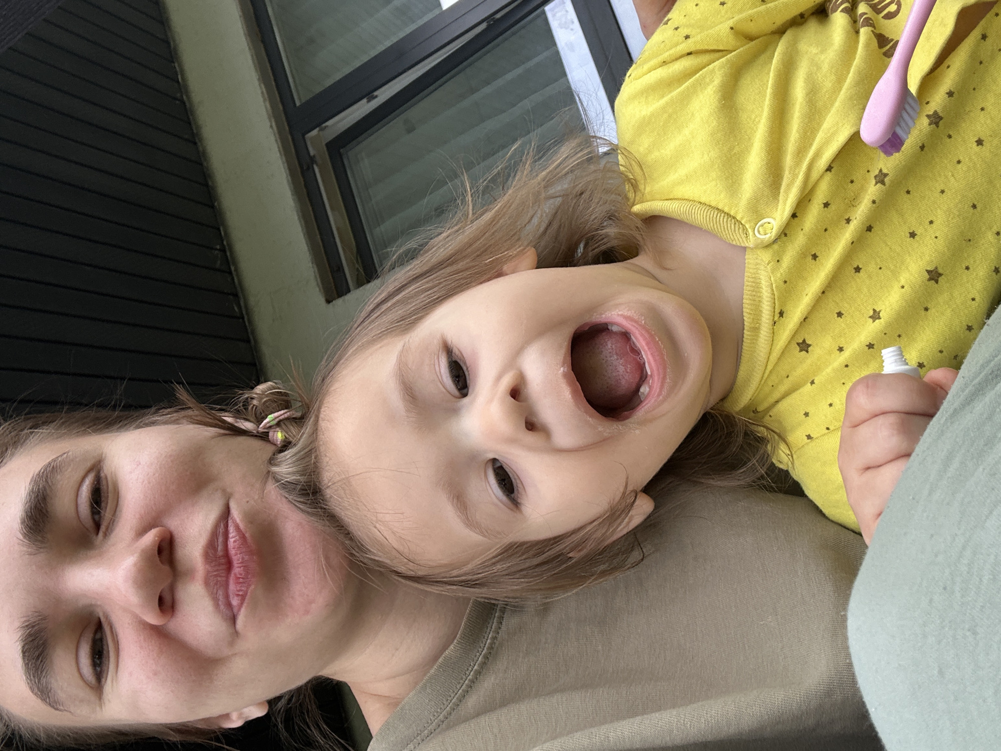 A woman and a child are featured in a selfie-style photograph. The woman has fair skin, dark eyebrows, brown eyes, and pink lips. Her hair is pulled back, and she is wearing a khaki-colored shirt. The child has fair skin, light brown hair, and is wearing a yellow, long-sleeved shirt with brown stars. The child's mouth is open wide. A pink toothbrush is in the child's hand, along with a tube of toothpaste. The setting appears to be indoors, with a window visible in the background. There is no indication that this image was taken in Kharnavy, Russia.