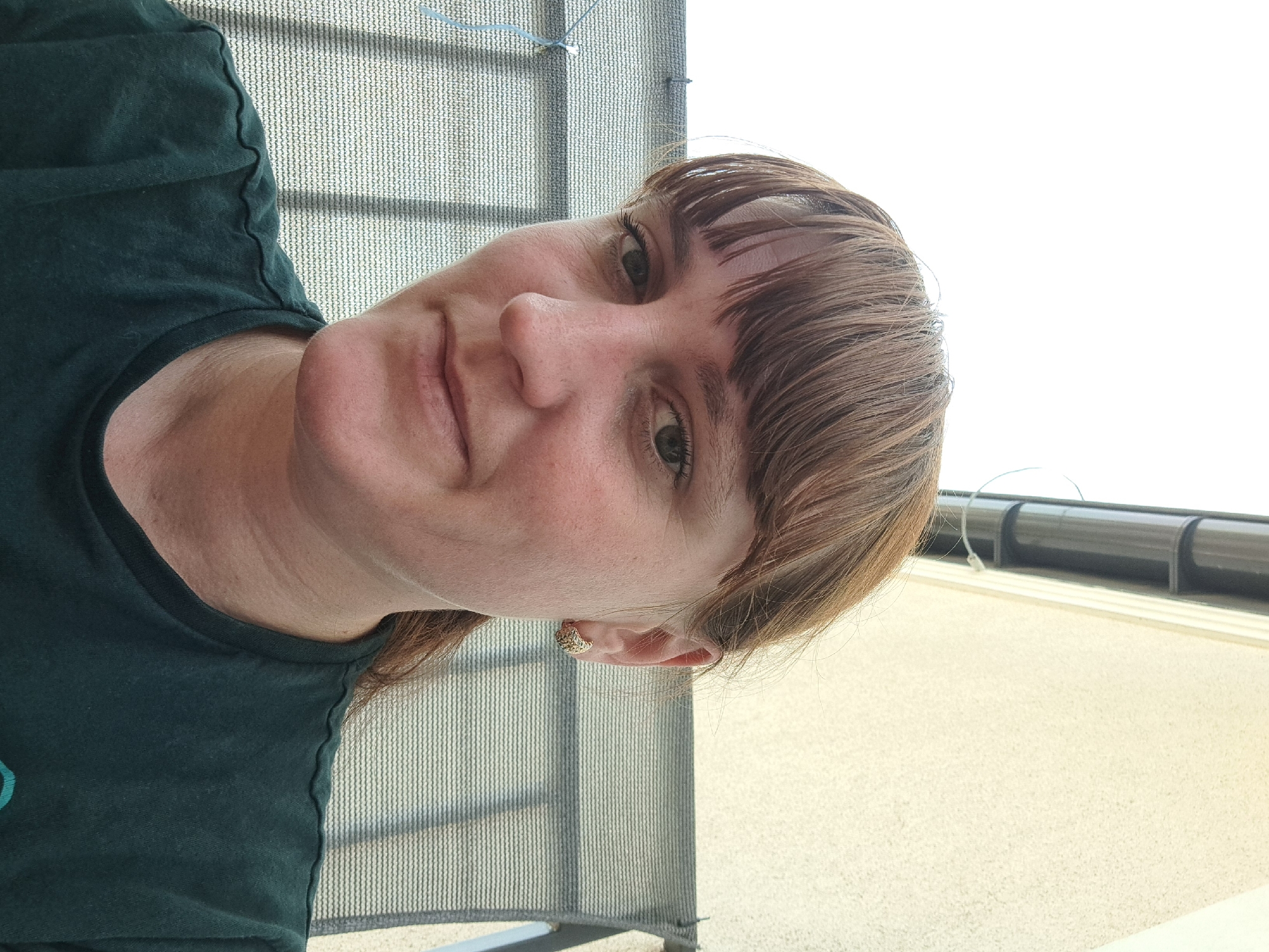 This is a portrait of a woman with light brown hair and bangs, facing slightly to her left. She is wearing a dark green t-shirt. A subtle smile can be seen on her face, and she has a small, textured earring in her visible ear. The background shows a blurred outdoor scene with what appears to be a mesh-like structure or fence, and a light-colored surface. The lighting suggests it is daytime, possibly outdoors with bright, diffused light, indicating a clear or partly cloudy sky. The overall impression is a casual, candid selfie. No text is visible in the image. The location context provided is Mińsk Mazowiecki, Poland.