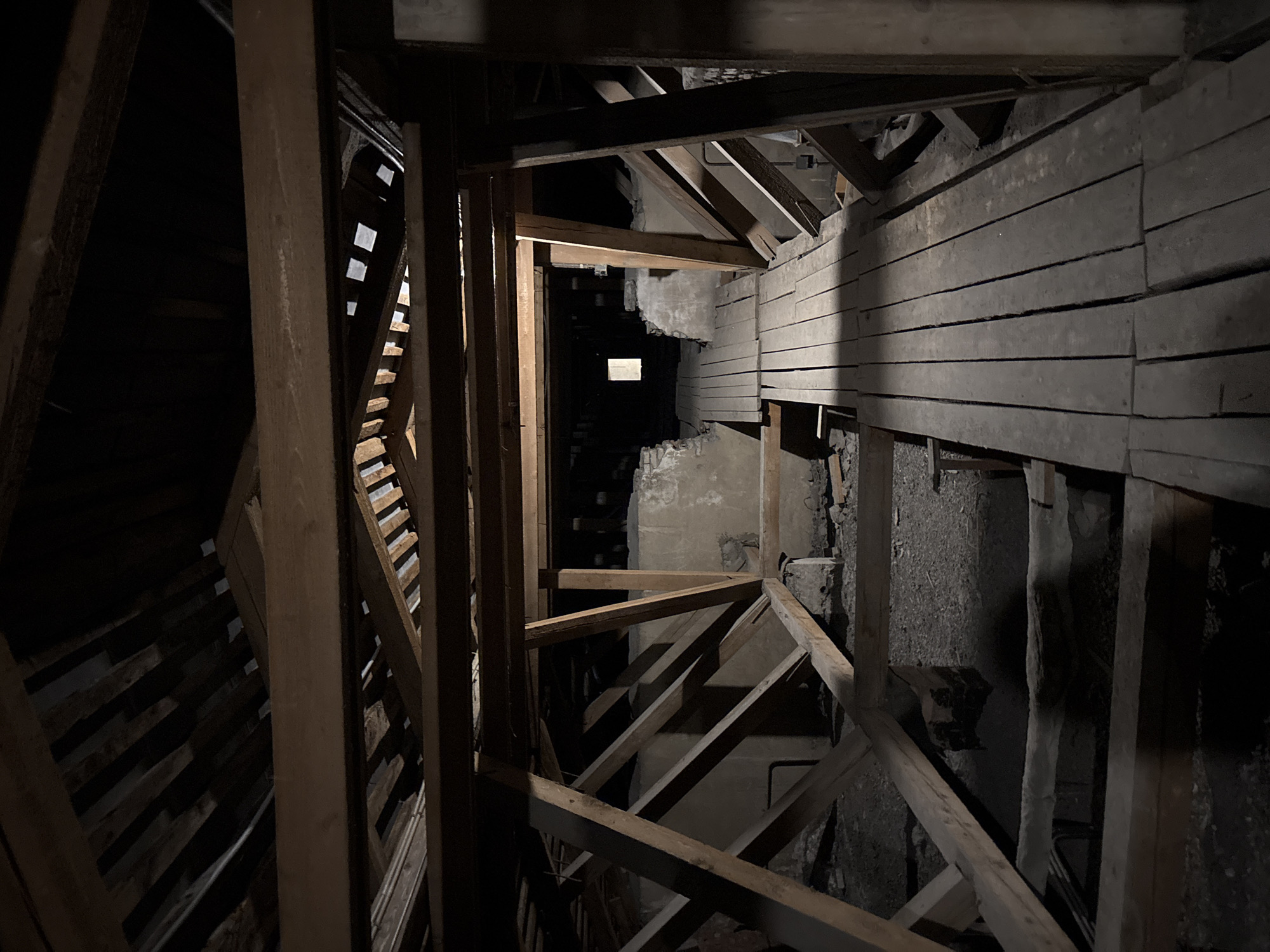 The image shows the interior of an attic in Krasnoye Selo, Russia. The attic has a wooden roof structure with exposed beams and rafters. A wooden walkway, constructed from planks, extends into the darkness. The walls appear to be made of concrete or stone, with sections of crumbling material. The overall impression is one of disuse and decay. There are no people or animals visible in the image. The scene is dimly lit, with a small square of light visible in the distance at the end of the walkway. The environment is dusty and appears to be undisturbed for some time.