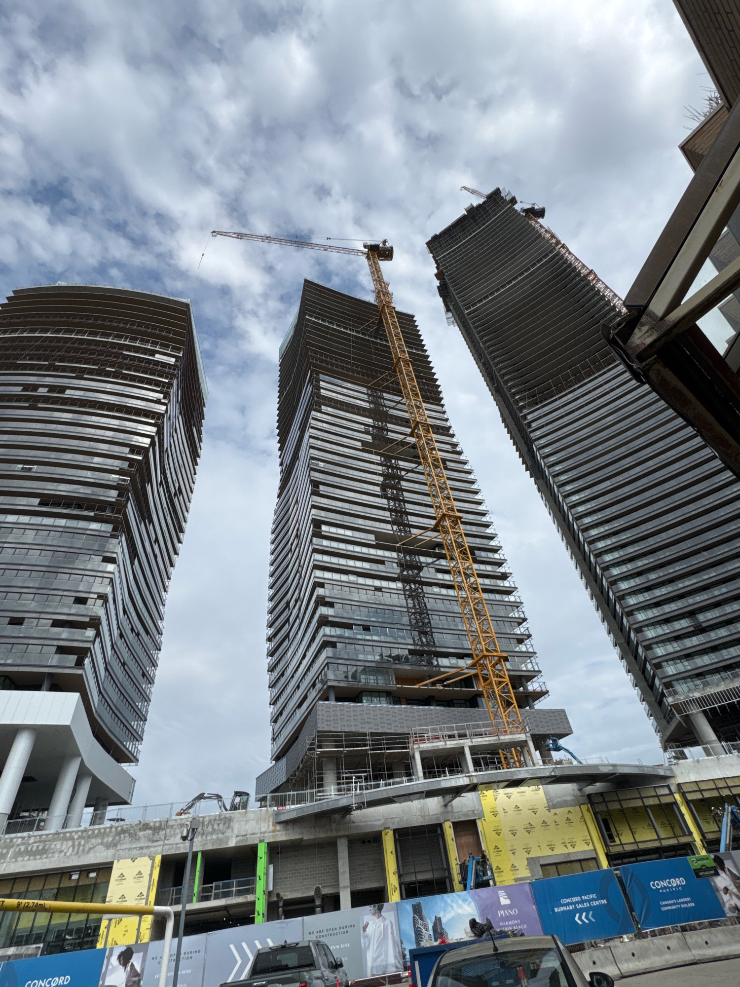 The image depicts a construction site in Burnaby, Canada, with three tall, modern skyscrapers under construction. Two of the buildings are in the foreground, their curved facades reflecting the cloudy sky. A large yellow construction crane is prominently positioned between the two foreground buildings, extending upwards. The third skyscraper is visible on the far right, also in its construction phase. The base of the buildings shows ongoing construction work, with scaffolding, barriers, and construction equipment like excavators. Signage for "CONCORD PACIFIC" and "PIANO" is visible at the lower portion of the image, indicating the developer and project name. The overall scene is one of urban development and progress.