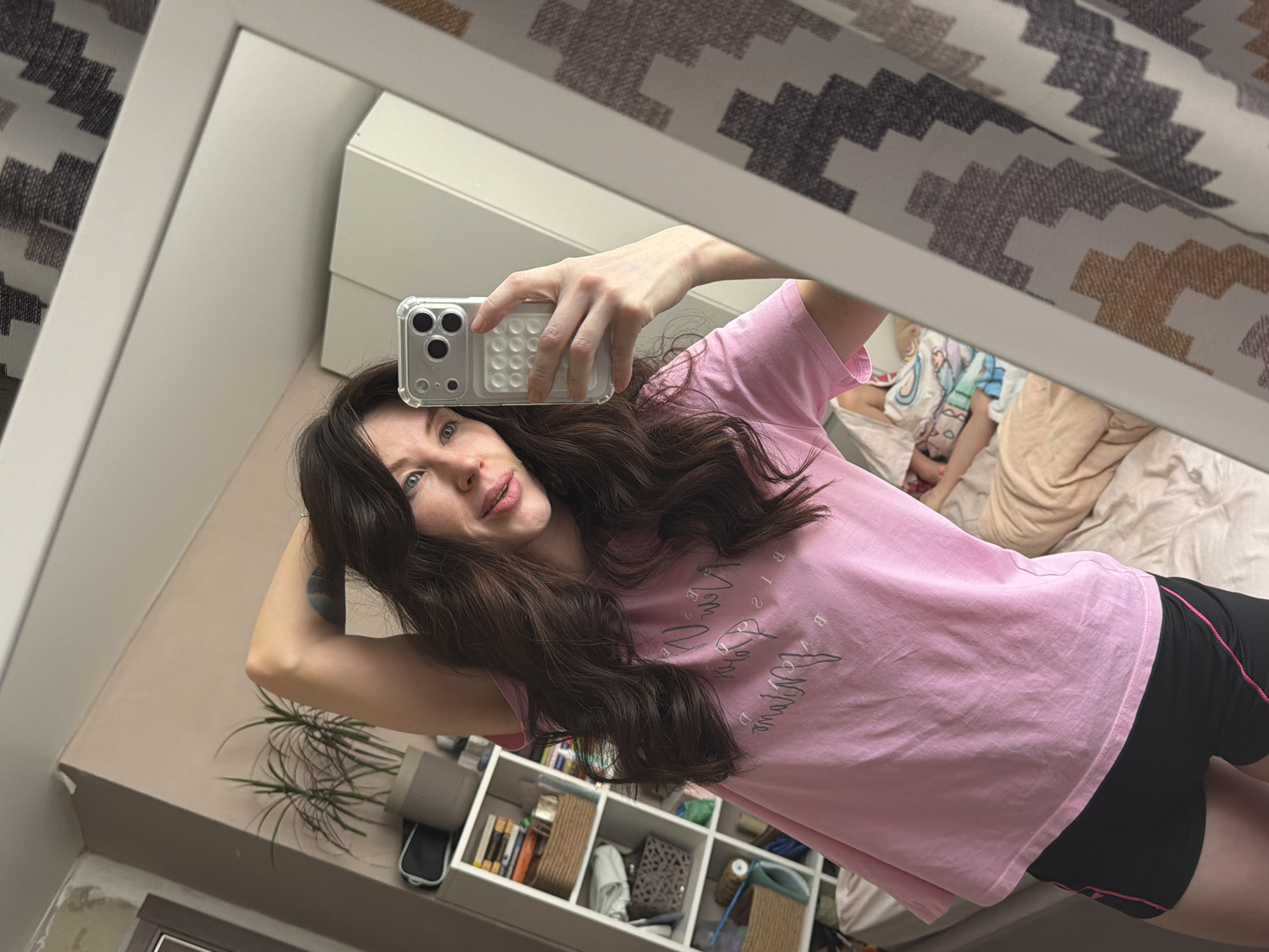 The image depicts an indoor residential setting, identified as being in Perm, Russia. The primary subject is an adult female with long, wavy dark brown hair and blue eyes, positioned in the foreground taking a mirror selfie. She wears a light pink short-sleeved t-shirt with dark, indistinct script on the front and black athletic shorts with pink piping. Her left arm is bent with the hand behind her head, revealing a dark, abstract tattoo on her upper left arm. She holds a silver-colored smartphone, featuring a clear case with a multi-circle pop-socket style grip and a triple-camera system, in her right hand. Her facial expression is a slight smile. Reflected in the background, a small child lies on a bed, partially covered by light-colored bedding, wearing a light-colored t-shirt with a printed character. The room features light beige walls, a white multi-shelf storage unit containing books and baskets, a potted plant with long green leaves, and a white wardrobe. A dark-framed door or window is partially visible on the left, and a patterned curtain with a zigzag design in brown, gray, and white tones is visible on the right.