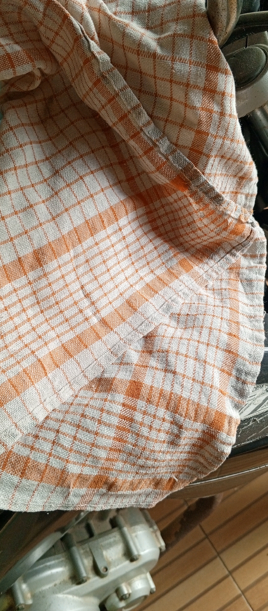 A close-up, overhead shot shows a crumpled, checkered dish towel with orange and white squares draped over a metallic object. The towel is folded and creased, with the weave of the fabric clearly visible. Part of a light-colored wooden floor and a dark surface are also visible in the background. The metallic object appears to be part of a machine or appliance, with visible bolts and a gray casing.