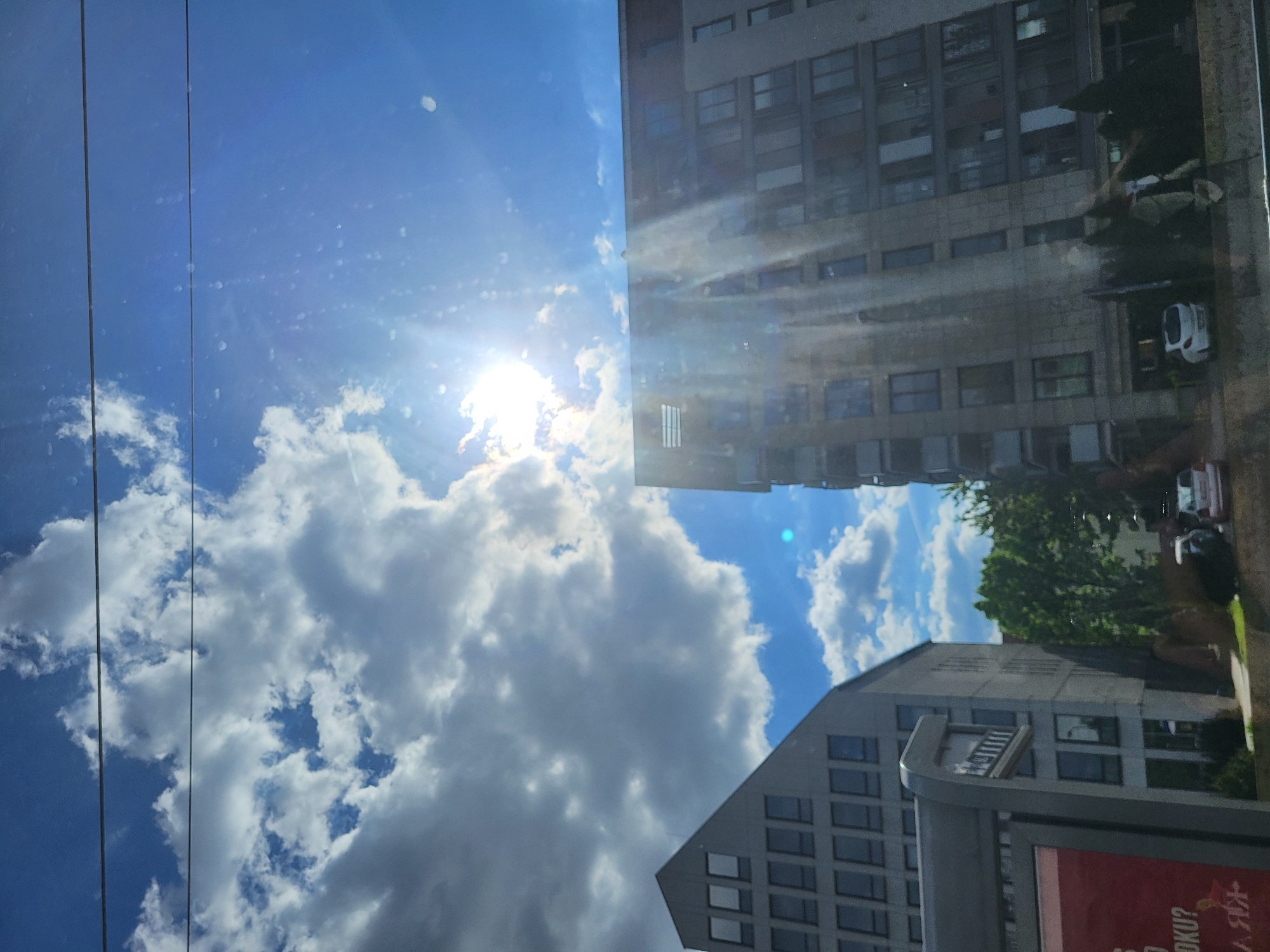 The image shows a view from within a vehicle, likely a bus or tram, looking up at buildings and the sky. The sky is predominantly blue with white cumulus clouds, and the sun is shining brightly. Two electrical wires run across the upper left of the frame. A tall, gray building with numerous windows occupies the upper right of the image. Reflections can be seen in the windows. A smaller, gray building is located in the lower-right corner. The view is partially obscured by a bus stop sign with a red advertisement panel. Vegetation (trees) are visible behind one of the buildings. Location is city of Unknown, Unknown.