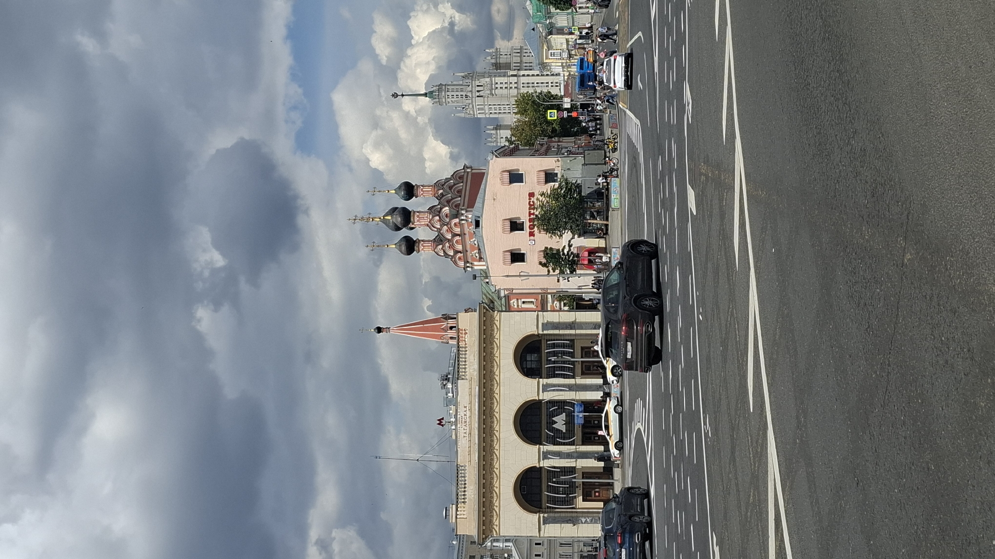 The image captures a street scene in Moscow, Russia, specifically near the Taganskaya metro station. In the foreground, a dark SUV is driving on a multi-lane road with white lane markings. To the left, the distinctive facade of the Taganskaya metro station is visible, featuring large arched entrances and the iconic Metro logo. Further to the right, a pink building with the "ROSTIC'S" sign is prominent, next to a red and pink church with multiple black domes. In the background, a tall, ornate white skyscraper, characteristic of Stalinist architecture, dominates the skyline. The sky is filled with dramatic, heavy grey clouds, suggesting overcast weather, with patches of blue sky peeking through. Several other cars and pedestrians are present in the mid-ground and background, indicating active city life.