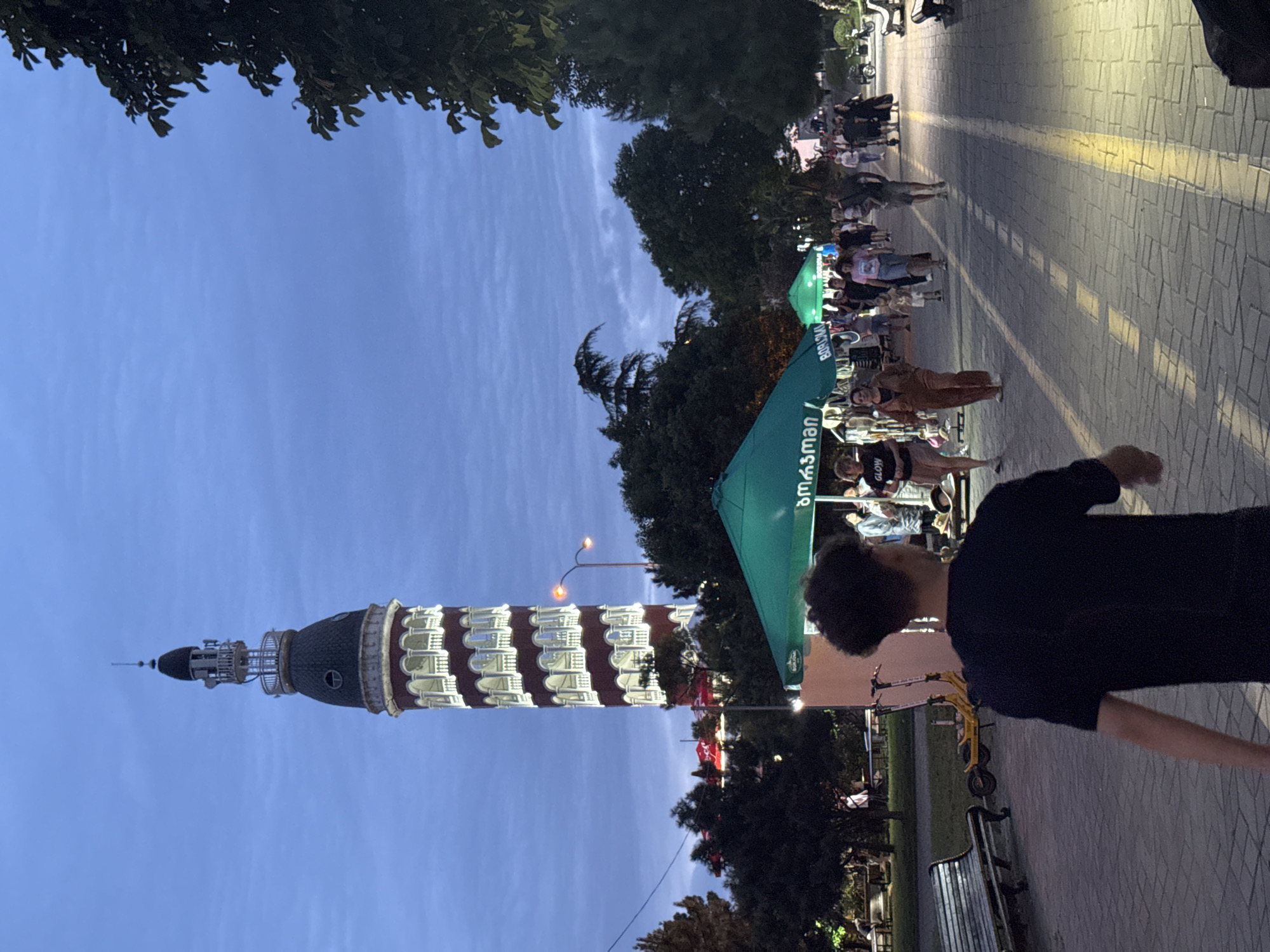 The image depicts a dusk scene in Batumi, Georgia, focusing on the Alphabet Tower and a pedestrian walkway lined with market stalls and people. The Alphabet Tower, a prominent landmark, stands tall in the background, illuminated against the twilight sky. In the foreground, a pathway paved with light-colored bricks stretches into the distance, casting long shadows from overhead lights.
Several market tents, predominantly green, are set up along the walkway, suggesting a temporary market or fair. People are seen walking along the path, browsing the stalls, and interacting with each other. Some individuals are captured in mid-stride, while others appear to be stationary, engaged in shopping or conversation. Among the crowd, a young person in a black t-shirt is walking away from the camera, their back to the viewer. Scooters and benches are also visible, indicating a public recreational area. The presence of trees and lush greenery suggests a park-like setting. The overall atmosphere is one of a lively evening gathering, common in tourist destinations like Batumi.