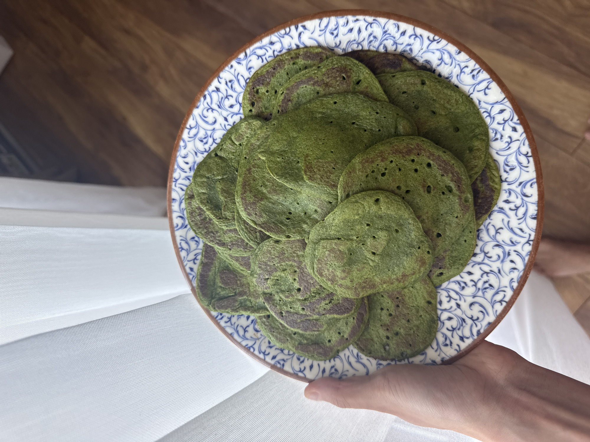 A hand holds a plate filled with a stack of green pancakes, presented on a decorative plate with a blue and white floral pattern, edged with a brown rim. The pancakes vary in size, displaying a speckled texture of green and darker brown hues, with small holes visible on their surfaces. The scene is set against a backdrop of light-colored, textured fabric, possibly a linen or cotton material, that appears to be a tablecloth or draping, and a wooden floor or surface in the background. The setting suggests an indoor environment.