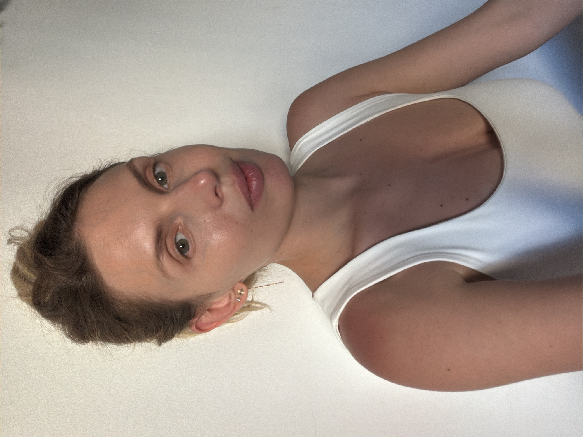 A fair-skinned woman with light brown and blonde hair is depicted in a close-up, indoor shot. She is wearing a white top. The background appears to be a plain white surface, possibly a wall. The location (Budva, Montenegro) cannot be confirmed from the image.