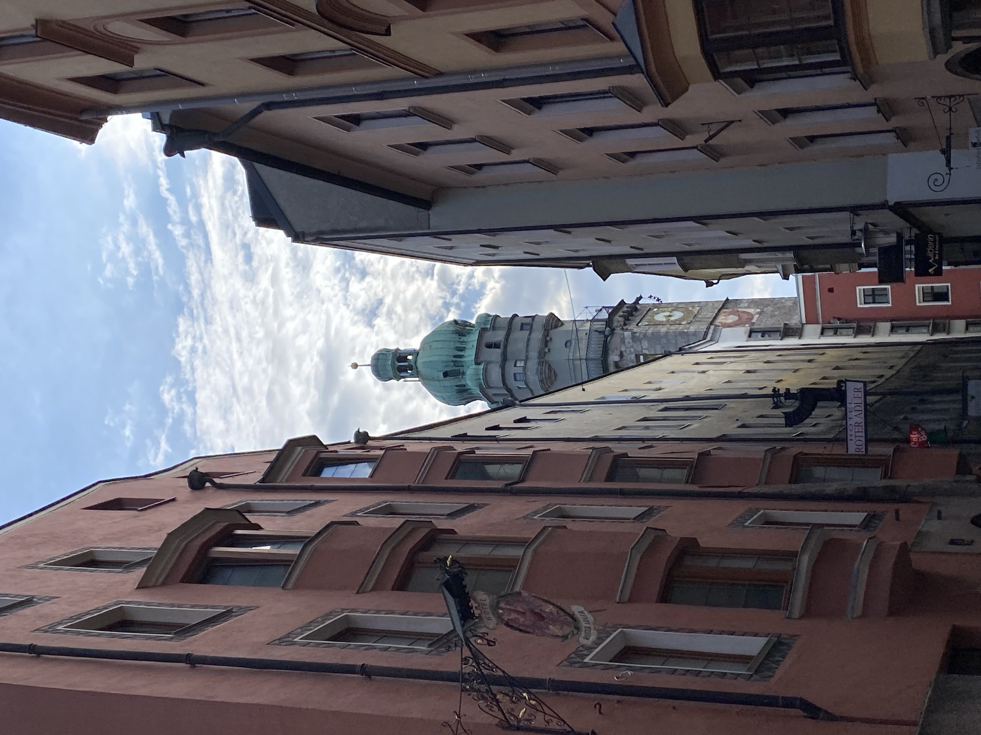 This is a low-angle, vertical shot of historic European architecture, likely in a city, with a prominent green domed tower visible in the distance. The buildings are several stories high, with multiple windows and ornate facades. The sky above is blue with scattered white clouds, suggesting fair weather, and the lighting indicates it is daytime, possibly late morning or afternoon.
The perspective is disorienting as the buildings appear to loom over the viewer, and the image is rotated, giving the impression of looking upwards from street level, or even from a tilted perspective. The architecture suggests a European location, and the context provided places it in Innsbruck, Austria.
Notable details include the stepped rooflines, the detailed window frames, and a hanging sign that is partially visible. The green tower in the background is a significant landmark, possibly a church or clock tower, with multiple levels and architectural ornamentation. There is a dark, metallic object hanging from one of the buildings, possibly an antique street lamp or a sign bracket. There is also a sign that reads "HOTEL ROTER ADLER". The image conveys a sense of historical depth and urban charm, with a slightly surreal or dramatic feel due to the unusual perspective and rotation.