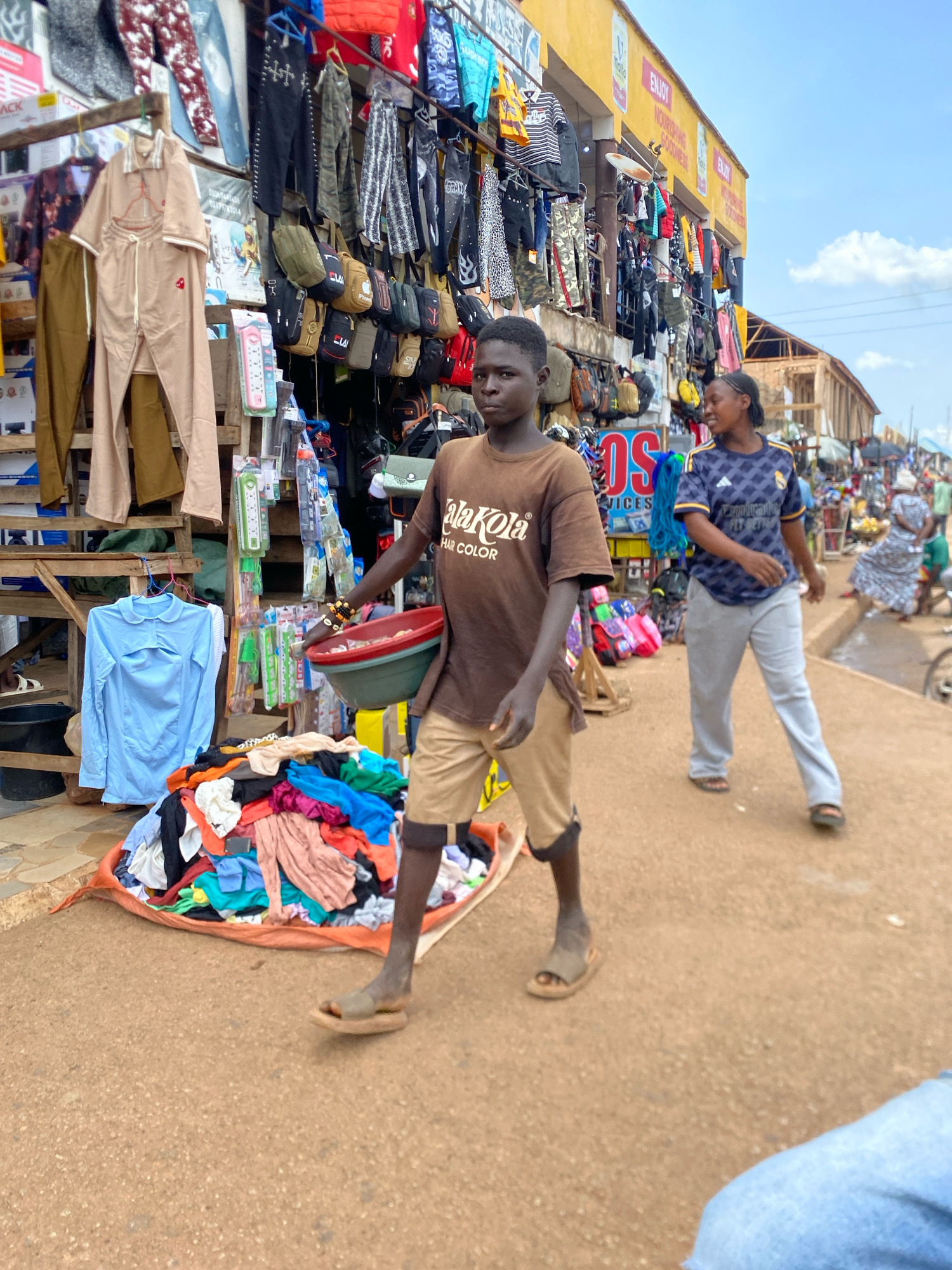 In the city of Bukuru, Nigeria, an outdoor market scene unfolds. In the foreground, a young Black male walks towards the right, wearing a brown t-shirt with "LaKola HAIR COLOR" printed on it, and tan shorts. He carries a red and green plastic bowl in his right hand, and his left arm is adorned with bracelets. He wears brown sandals. To his right, a young Black female walks in the same direction. She wears a dark blue and white patterned t-shirt with a logo that appears to be "Adidas" and gray sweatpants, also wearing sandals. Behind them, a stall displays a variety of clothing and accessories. Rows of pants, shirts, and bags are hung on a wooden rack. In the lower left corner, a pile of colorful clothes is spread out on an orange sheet. Further down the street, other market stalls and people are visible, with some buildings in the background. The ground is a sandy, reddish-brown color, and the sky is partly cloudy.