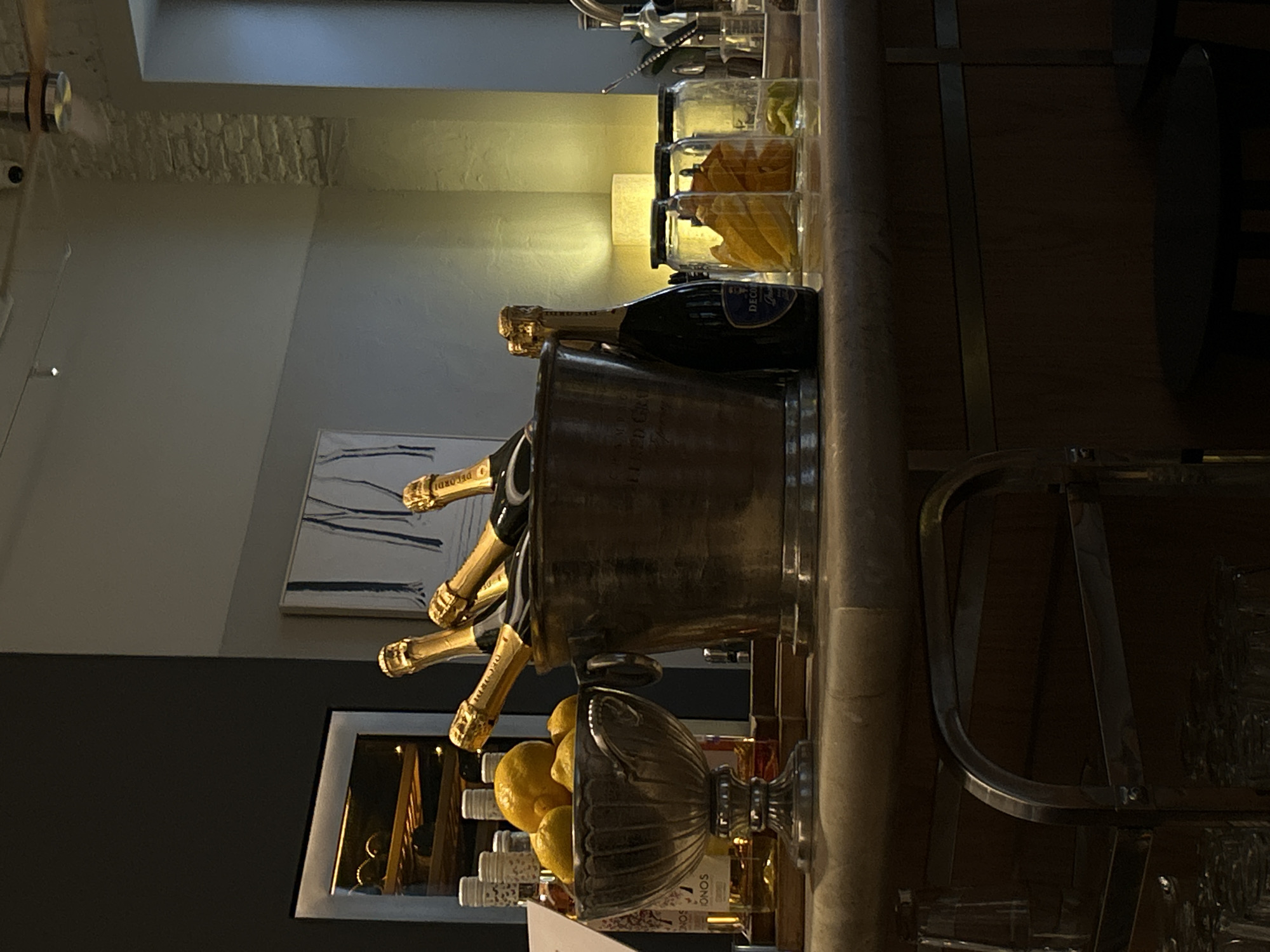 The image captures a well-appointed bar counter, likely in a restaurant or hotel, with a focus on beverages and garnishes. The scene is illuminated by warm, artificial light, suggesting evening hours or an indoor setting.
In the center, a large, metallic ice bucket holds several bottles of champagne, with golden foils and dark labels prominently displayed. One bottle, labeled "DECORDI," stands outside the bucket on the dark wooden counter. To the right of the champagne, several clear jars contain dried fruit slices, including what appear to be oranges and limes, serving as cocktail garnishes. Behind these jars, a soft, warm glow emanates from a lamp, highlighting a section of the wall above.
Further left on the counter, a decorative silver bowl or urn is filled with fresh lemons. Behind it, various bottles of spirits are partially visible, one clearly marked "SONOS." The counter is dark wood, complementing the overall sophisticated ambiance.
The background reveals architectural details. Above the counter, an abstract painting with blue and white strokes hangs on a light grey wall. Higher up, a section of exposed light-colored brickwork is visible, adding a rustic or industrial-chic touch. Structural elements, such as a metal bar frame and part of a column, are visible on the right side. Stacked glassware is partially seen beneath the counter on the far right. No people are present, giving the impression of a prepared, awaiting space.