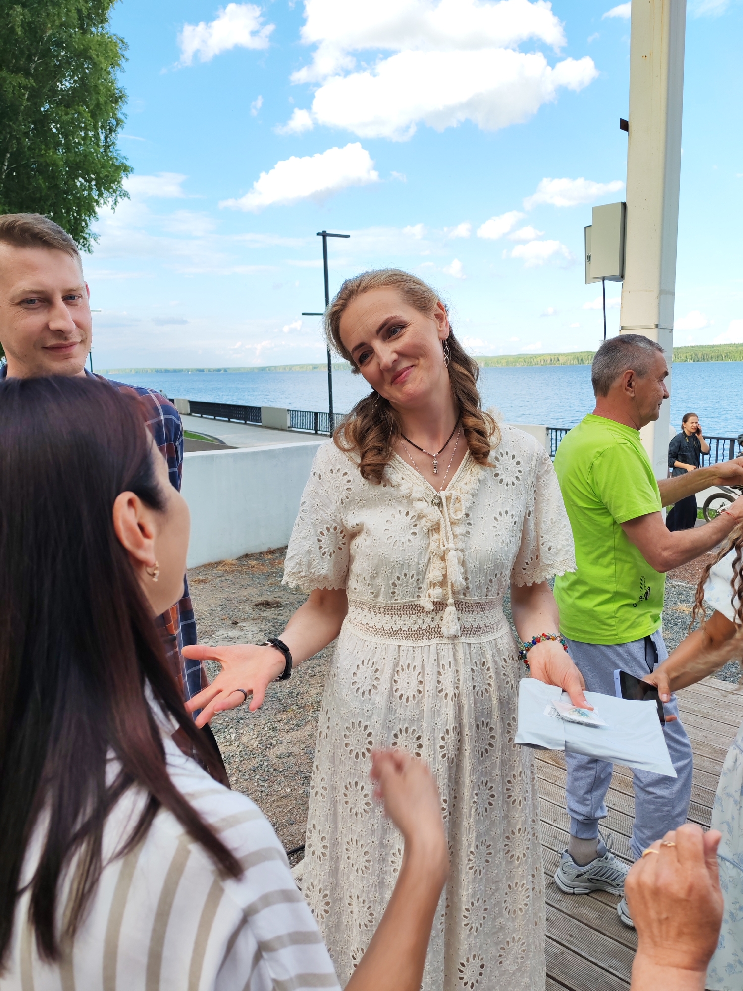 In Chaikovsky, Russia, near the Kama River embankment, a group of people are gathered outdoors. A woman in a white lace dress is speaking with a woman in a striped shirt, gesturing with her hands. Behind them, a man in a plaid shirt looks at the camera, and to the right, a man in a green shirt holds a phone. In the background, a body of water and trees can be seen, under a sky with white clouds. The ground is covered with gravel and wooden decking.