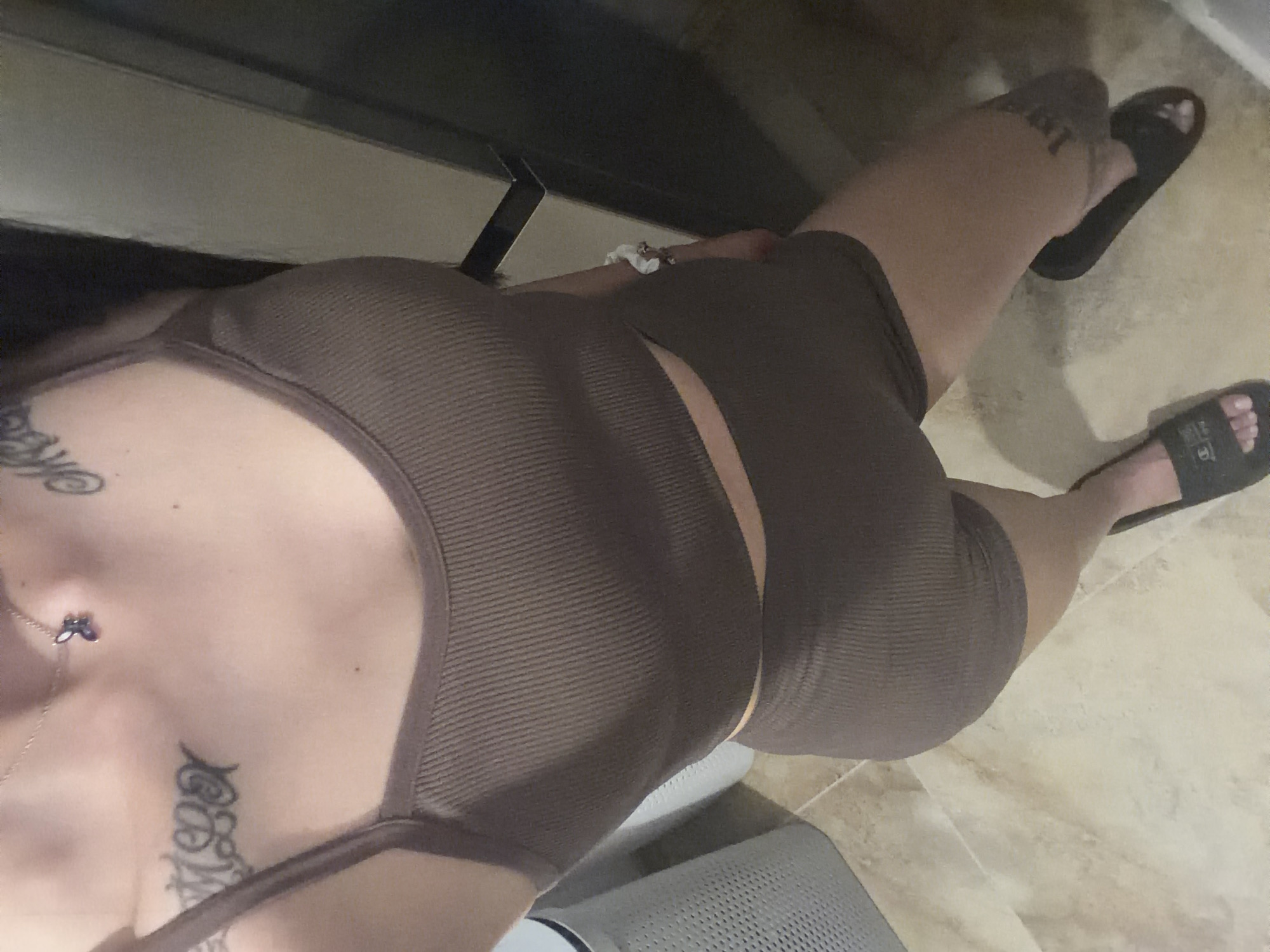 The image shows a close-up of a person wearing a brown ribbed two-piece outfit consisting of a crop top and shorts. Tattoos are visible on the person's chest, upper arm, and thigh. The person is wearing black slides. The background includes a tiled floor, a grey laundry basket, and a dark surface that appears to be a refrigerator. The image is taken from a high angle, looking down at the person. There is no information in the image to suggest a location in Plovdiv, Bulgaria.