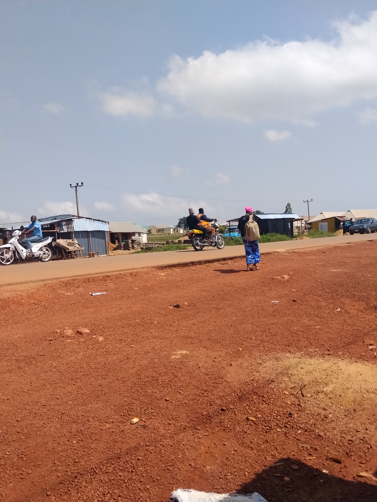 In the city of Durumi, Nigeria, on a dirt road, a man rides a white motorcycle. Further down the road, two people ride a yellow motorcycle, and a person with a pink hat and a backpack walks away from the camera. In the background, there are several buildings, some made of corrugated metal, and a parked car. The sky is blue with white clouds.