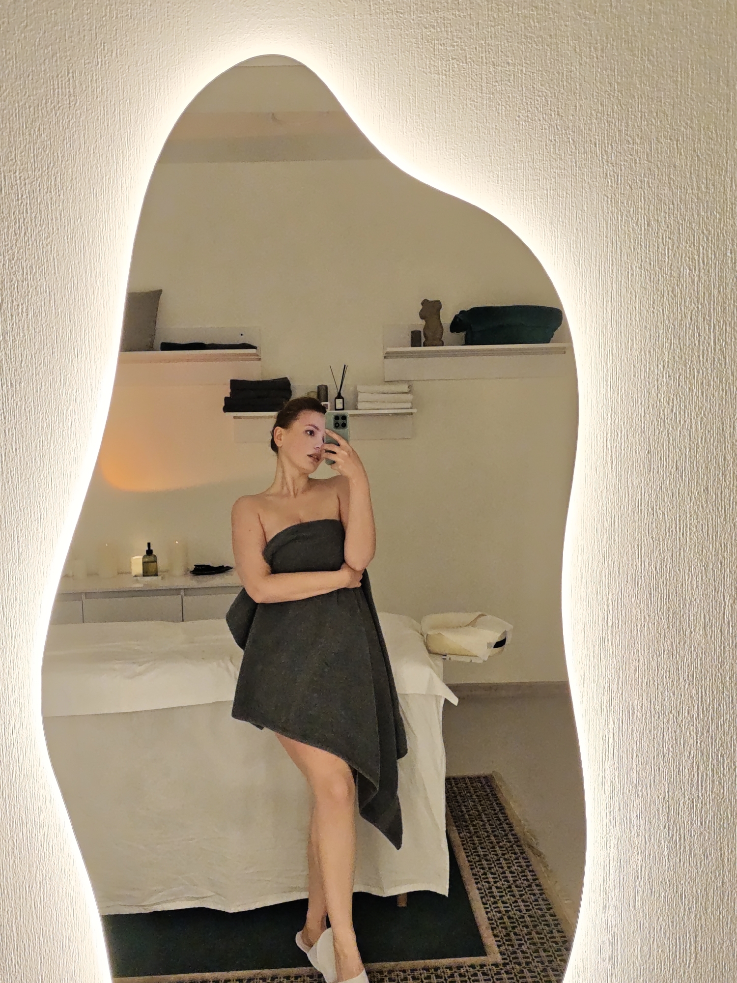 A woman wrapped in a gray towel stands in a room, as reflected in a uniquely shaped mirror with built-in lighting along its edges. The room appears to be a spa or treatment room, featuring a massage table covered with white linens. Two shelves are visible on the wall behind the table, holding items like towels, candles, and decorative objects. The flooring includes a dark green rug partially covered by a patterned carpet. The woman is holding a smartphone, presumably taking a selfie. It is not possible to confirm the location as Vinnytsia, Ukraine from the provided image.