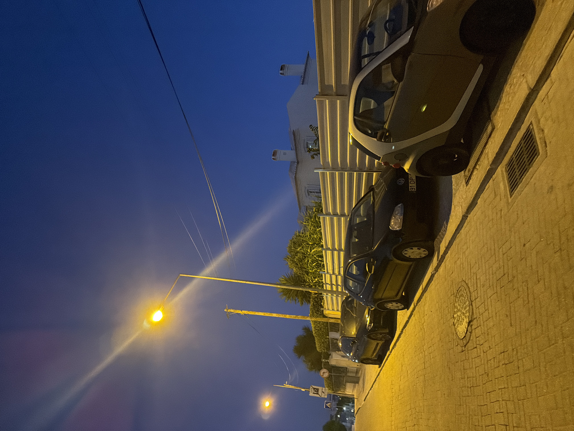 This is a nighttime, eye-level, outdoor, medium shot of a street scene in Cascais, Portugal. The scene is illuminated by a streetlamp and the ambient light of dusk. The sky is a deep blue, transitioning to a lighter hue near the horizon. A streetlamp with a bright yellow light is prominent on the left side of the image. Electrical wires stretch across the sky.
Parked on the right side of the street are several cars. Closest to the viewer is a small, two-door smart car with a white roof and light gray body. To its left, a black Volkswagen Golf is parked, its license plate visible as "91-DP". Further down the street, another dark-colored car, possibly a BMW, is parked.
The street is paved with light-colored bricks. A manhole cover is visible in the foreground, centered on the brick pavement. A metal fence runs along the right side of the street, separating it from a property with a white building and two chimneys visible in the background. Lush green bushes and trees are planted behind the fence. A traffic sign with a directional arrow is visible on the left side of the street, near a pedestrian crossing sign. A yellow lamppost stands between the cars and the fence. The overall scene depicts a quiet, residential street at night.