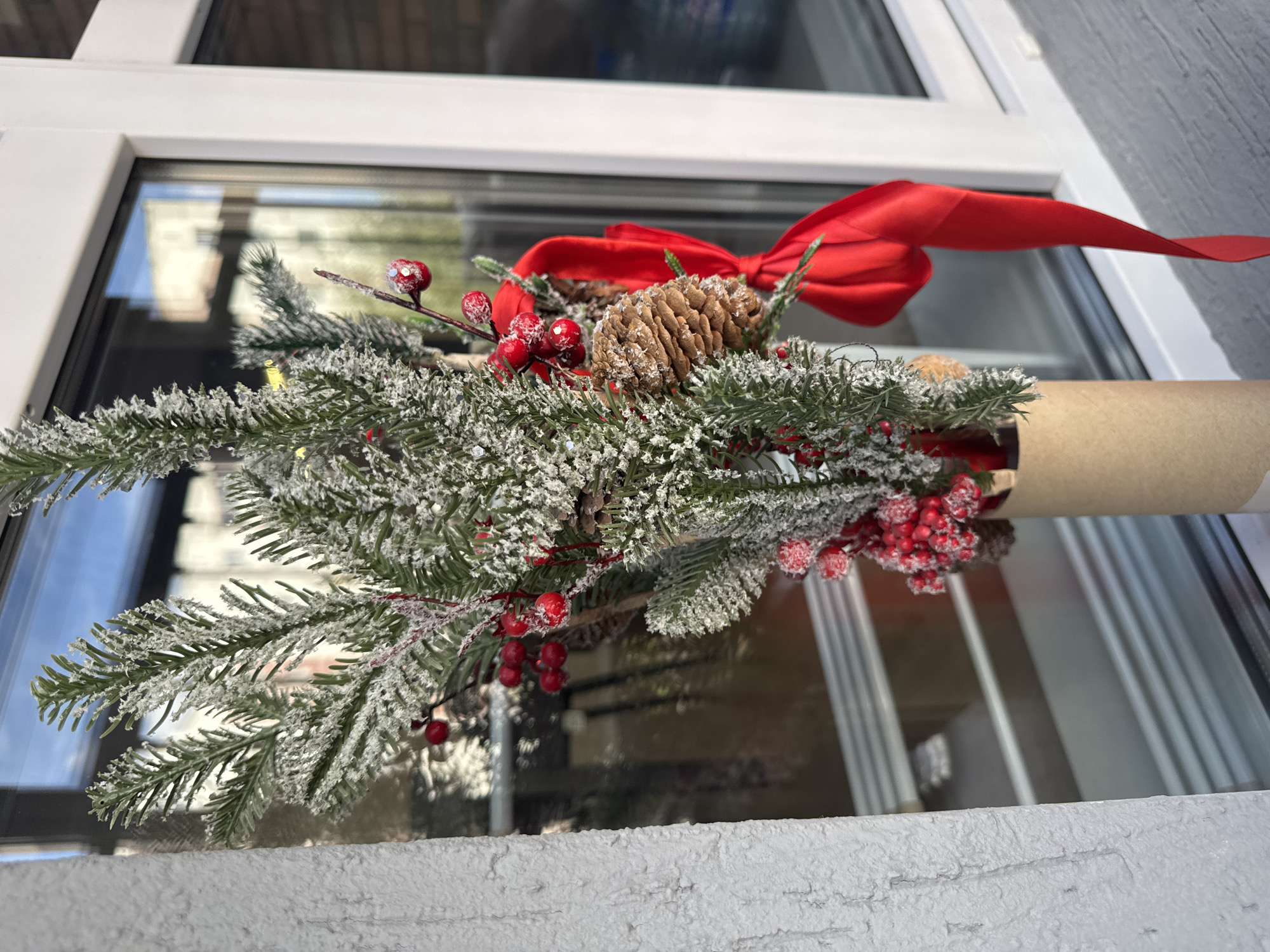 This is a medium shot, eye-level, outdoor, daylight photograph of a Christmas-themed decoration. The decoration is in a cylindrical beige holder and consists of artificial evergreen branches dusted with white material to simulate snow. Accompanying the branches are several clusters of small red berries and a few pinecones. A bright red ribbon is tied in a bow and trails down from the arrangement. The decoration is placed on a windowsill, with a window and its frame visible to the right. Reflections of buildings and the sky are visible in the window glass. A textured, white wall is visible to the left of the decoration. The scene appears to be in the city of Perm, Russia.