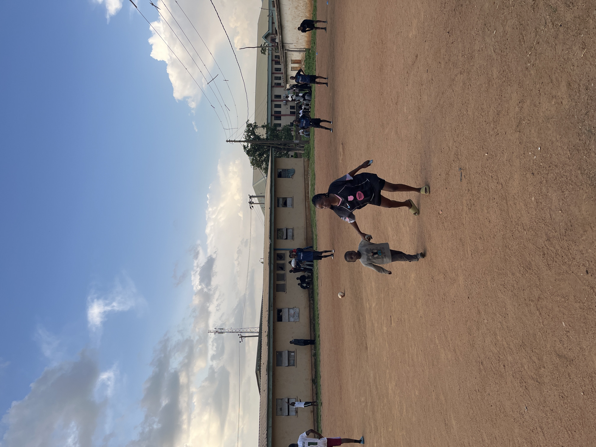The image shows a group of people, predominantly young men and boys, on a dusty field in Jos, Nigeria. The setting appears to be an outdoor sports area adjacent to a row of low-rise buildings, possibly educational or residential. The activity taking place is likely a casual game of football or a practice session, as a football is visible on the ground.
Several individuals are in motion, suggesting active participation. One person in the foreground, wearing a black and pink jersey, is in mid-stride, facing away from the camera. Another person in a grey t-shirt and dark shorts is also engaged in an activity, possibly receiving or kicking the ball. In the background, a larger group of people, many wearing dark jerseys with blue accents, are positioned along the side of the field or in the buildings.
The time of day appears to be late afternoon, indicated by the warm, soft lighting and the shadows cast on the ground. The weather is clear with some scattered clouds in a bright blue sky. The location cues, such as the architecture of the buildings and the general environment, are consistent with a West African setting, specifically Jos, Nigeria. There are power lines visible overhead.
No specific emotions are clearly discernible from the image due to the distance and the dynamic nature of the activity. The overall impression is one of energetic, informal recreation. There is no visible text in the image.