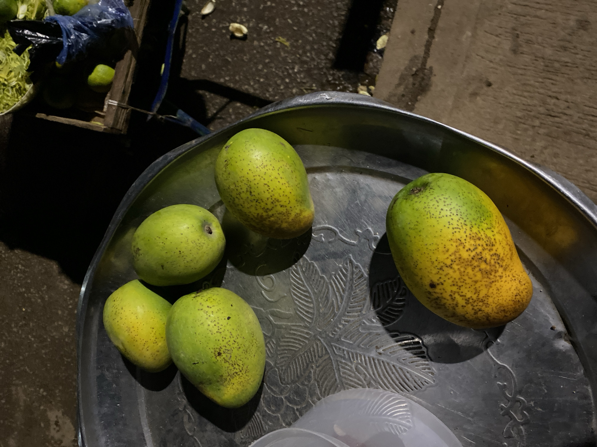 The image presents an overhead view of five green and yellow mangoes arranged on a dark, metallic tray, likely at a street market in Bukuru, Nigeria, during the evening or night. The mangoes are in various stages of ripening, displaying a mix of vibrant green and yellow hues, and their skins are speckled with small, dark spots. The round tray features an intricate embossed pattern of leaves and other decorative elements.
In the lower foreground, a section of a translucent white plastic container, also with a textured design, is visible beneath the tray. The background is dimly lit by artificial light sources, casting long shadows and highlighting the texture of the ground, which appears to be asphalt or concrete with scattered light-colored debris. To the left, a blue plastic bag is seen next to what appears to be a wooden crate or stand, behind which a container holds lush green, leafy produce. The low light conditions strongly indicate that the scene is set after dusk. No people or visible text are present in the image. The arrangement of the mangoes suggests they are being displayed for sale.