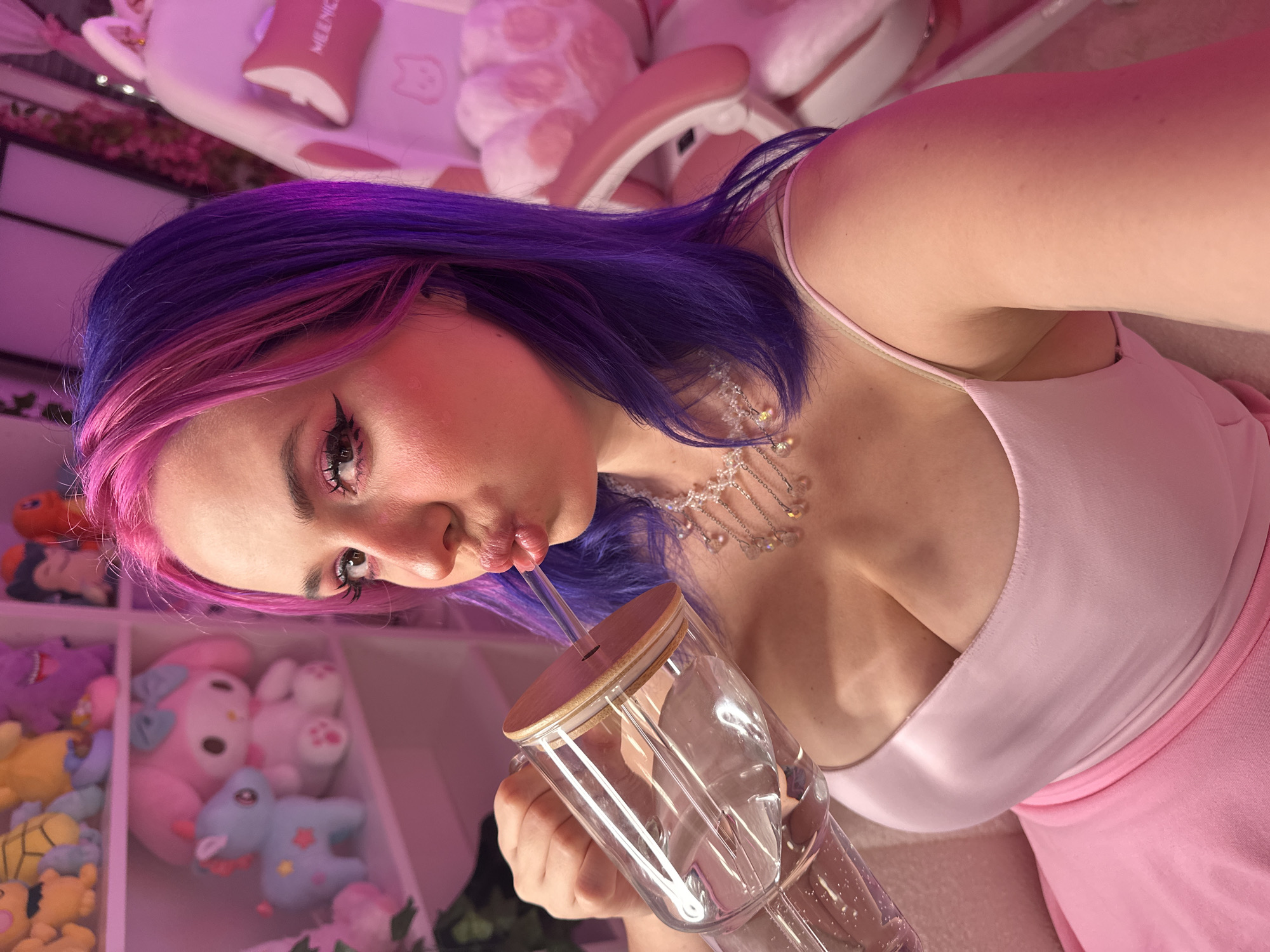 A woman with pink and purple hair is drinking from a glass mug with a straw. She is wearing a pink dress and a necklace. The background includes shelves filled with stuffed animals and a pink gaming chair. There is no indication that this image was taken in Oviedo, Spain.