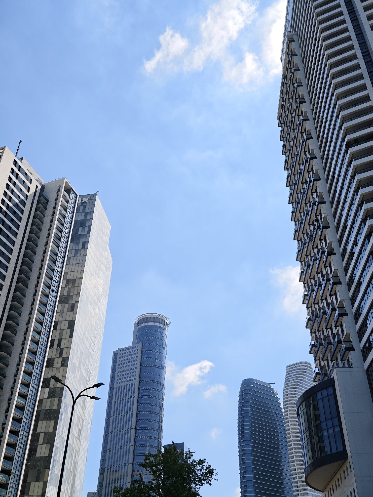 The photograph captures a low-angle view of modern skyscrapers against a clear blue sky with scattered white clouds. The buildings are of various architectural designs, with glass facades, balconies, and distinct geometric shapes.
On the left, a tall, light-colored building with repeating horizontal lines and dark window sections dominates the frame. To its right, a taller, slimmer building with a reflective glass facade and the letters "DN" visible near the top. Further into the background, a cylindrical skyscraper with a light blue hue and a helipad at its crown stands out. To the right of this, a curved, dark blue skyscraper with horizontal white banding is visible, followed by a white, stepped skyscraper. On the far right, a portion of another tall building with a patterned facade and protruding balconies is shown.
A street lamp with multiple light fixtures stands in the foreground, to the left of the buildings. Lush green trees are visible at the bottom of the frame, partially obscuring some of the lower sections of the buildings.
The image was taken in Givatayim, Israel, and showcases the contemporary urban landscape of the city. No people are visible in the image. The scene is static, focusing on the architecture and the sky.
