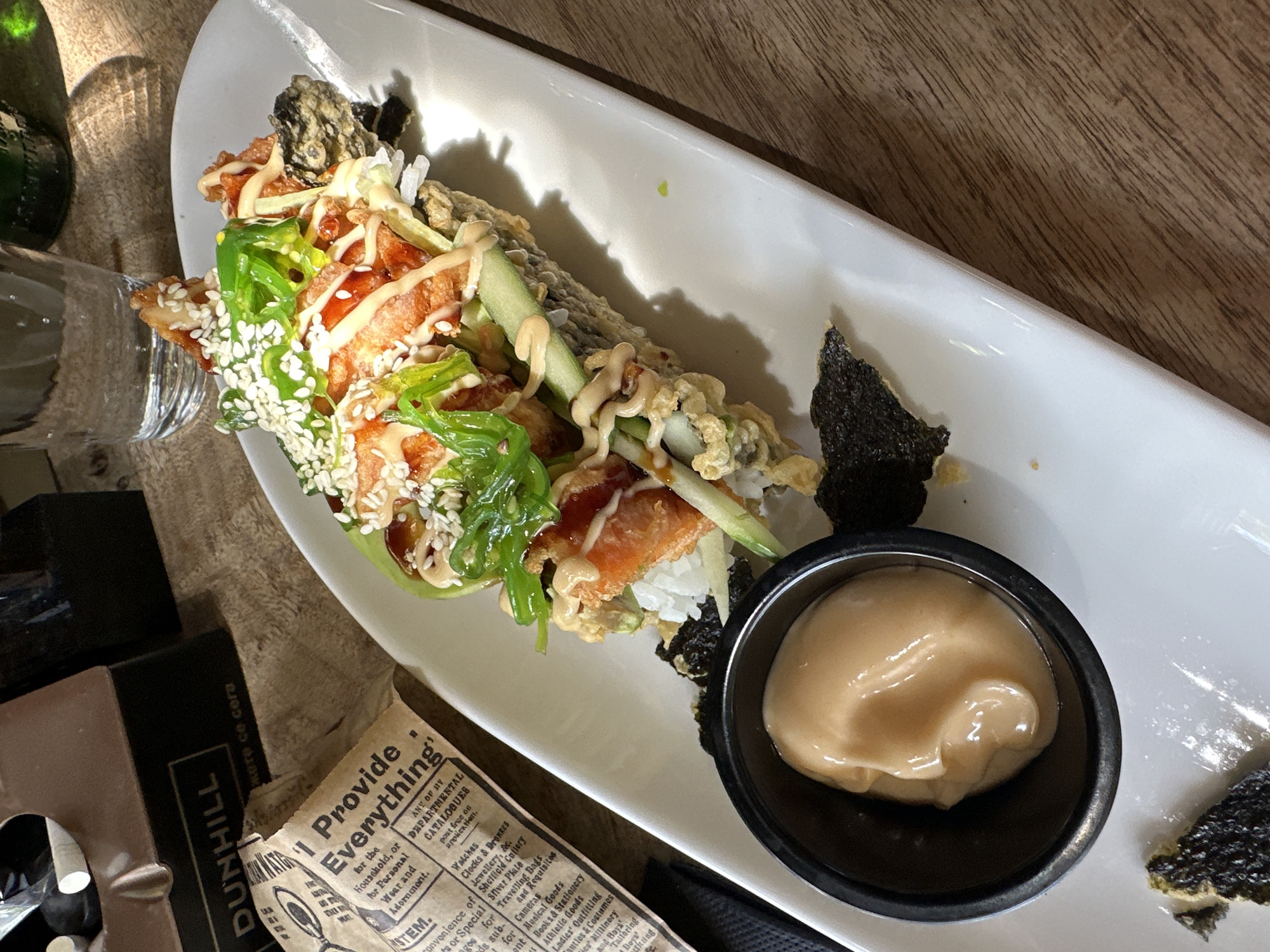 A close-up shot captures a plate of sushi on a wooden table. The sushi roll is presented on a white rectangular plate and appears to be deep-fried, topped with what looks like seaweed salad, cucumber slices, and a drizzle of sauce. A small black bowl containing a creamy dipping sauce sits to the left of the sushi. In the background, out of focus, are a glass of water, a dark object with the text "DUNHILL" visible, and a hint of a green bottle. The lighting suggests it is daytime, with a shadow cast on the plate and table from an unseen light source. The overall scene is a still life focused on the food presentation. The image was taken in Sofia, Bulgaria.