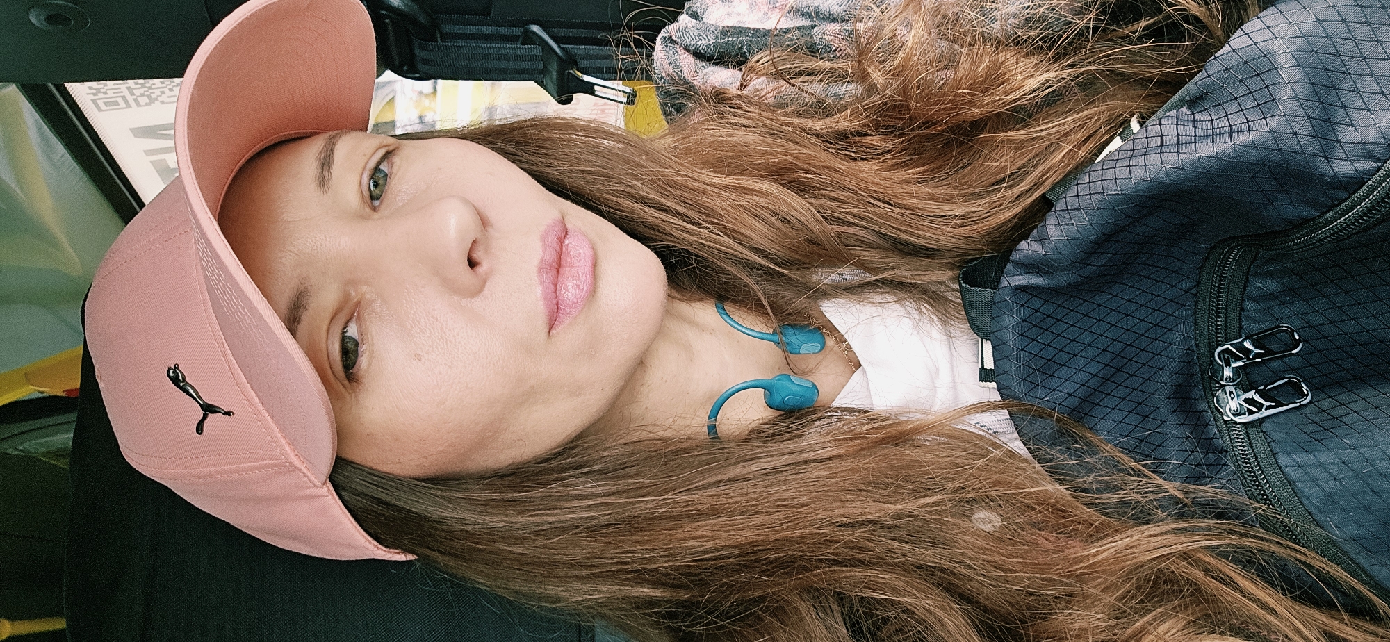 A close-up, slightly high-angle, selfie-style portrait shows a woman wearing a pink baseball cap with a black Puma logo. She has long, brown hair, light skin, and is wearing blue earbuds around her neck. She is also wearing a white shirt and a plaid jacket, partially visible. In the foreground, a dark blue backpack with a diamond pattern and two silver zipper pulls is visible. The background is out of focus and appears to be the interior of a vehicle, with a hint of yellow and a QR code visible on what might be a sticker. The specific location within the city of Vitsebsk, Belarus, cannot be determined from the image alone.