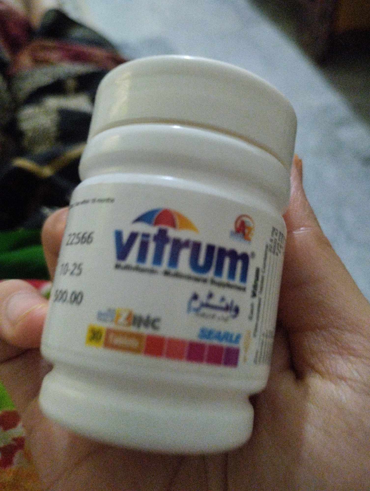 The image shows a close-up of a white plastic bottle of "Vitrum" multivitamins. The bottle is held in a person's hand, with the background out of focus and appearing to be an indoor setting with patterned fabric.
On the bottle, the brand name "vitrum" is prominently displayed in large blue letters with a colorful arc graphic above it. Below "vitrum", the text "Multivitamins. Multimineral Supplement" is visible. There is also text in Arabic script, which appears to be "وانشرم".
To the left of the brand name, there are numbers and text: "22566", "10-25", and "500.00". Towards the bottom of the bottle, the word "ZINC" is visible with a colorful bar below it featuring squares of different colors. The name "SEARLE" is also visible towards the bottom.
The lighting suggests it might be indoors, possibly during the day or with artificial light. No specific time of day or weather conditions can be determined. The focus is on the product, and the person's hand is visible holding it, but their face or any other identifiable features are not shown. The overall impression is a simple product shot.
