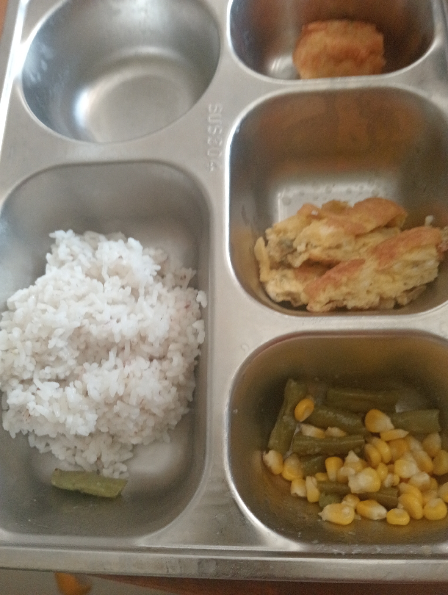 The image shows a close-up, overhead view of a metal lunch tray with three compartments filled with food. The top left compartment is empty. The top right compartment contains a single, round, golden-brown breaded item. The middle compartment is filled with white rice. The bottom compartment contains green beans and yellow corn kernels. A thin, green slice, possibly a piece of cucumber or pickle, is visible at the bottom of the rice compartment. The background is out of focus and appears to be a table surface. The location in Wanasalam, Indonesia, is not visually represented in the image.