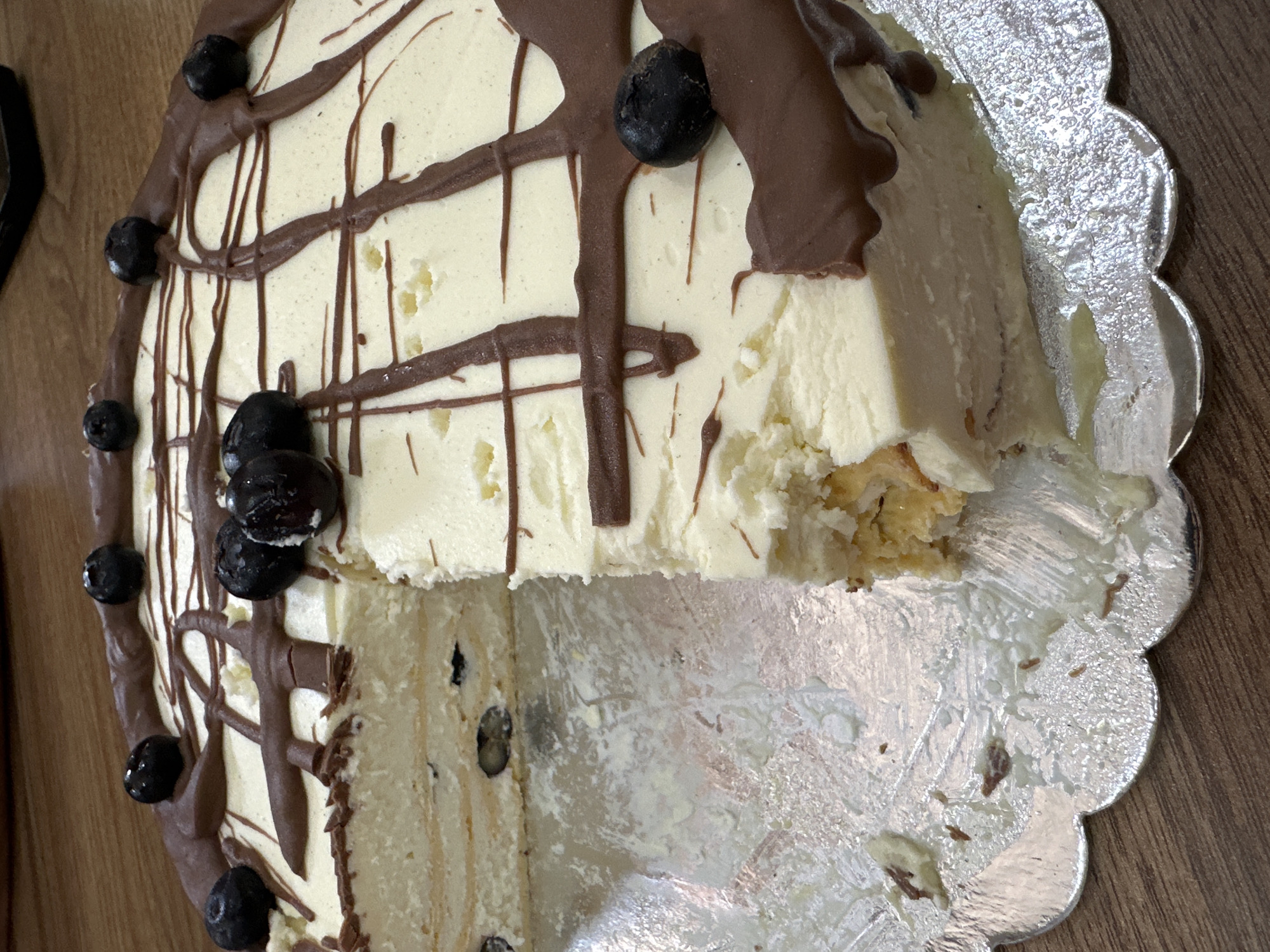 A partially consumed round cake rests on a decorative silver cake stand on a wooden surface. The cake is frosted with a light-colored cream, drizzled with brown chocolate, and adorned with fresh blueberries. A wedge has been cut from the cake, exposing the layered interior. The remnants of cake and cream are visible on the cake stand. The image was taken in Lehchevo, Bulgaria.