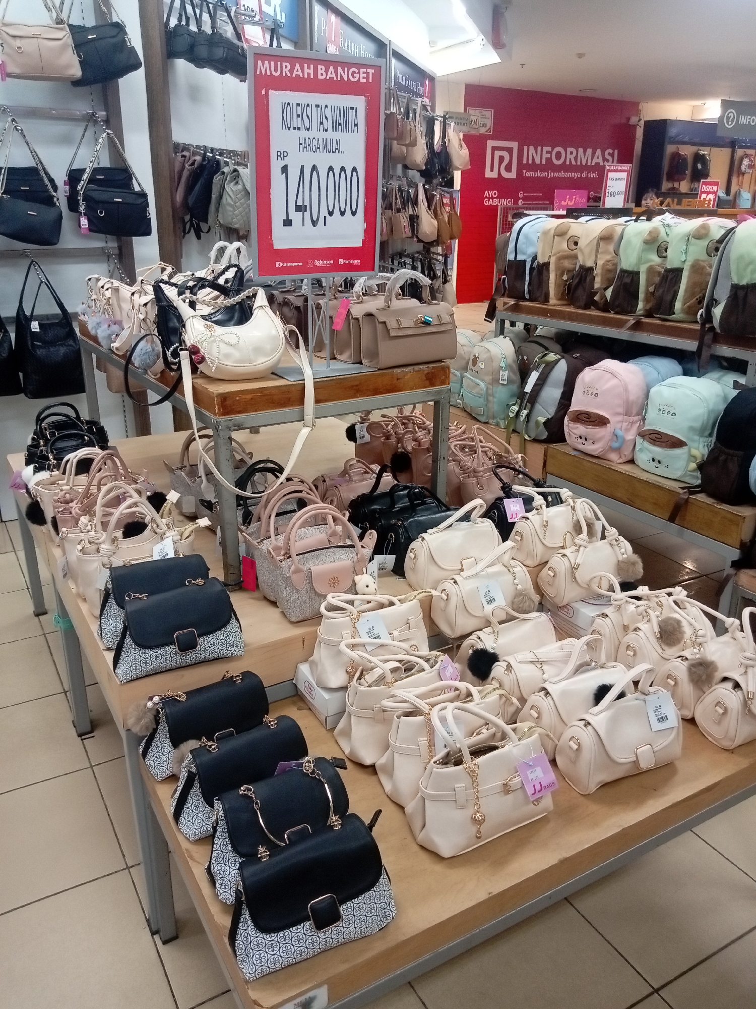 The image depicts a retail display of handbags and backpacks in what appears to be a department store in Batam, Indonesia, likely within the Robinson department store chain, as indicated by signage. A prominent red sign displays "MURAH BANGET" (Very Cheap) and "KOLEKSI TAS WANITA HARGA MULAI.. RP 140.000" (Women's Bag Collection Starting Price.. RP 140,000).
The foreground and midground are filled with various handbags, mostly in neutral tones like cream, beige, and black. Many of these bags are smaller, shoulder bags, or crossbody styles, some adorned with decorative charms like pom-poms or keychains. There are also several structured tote bags and clutches. Some of the handbags are displayed on wooden shelves, while others are presented on metal racks. In the background, rows of backpacks are visible, featuring colorful designs and cartoonish prints, suggesting they are intended for a younger demographic. The store environment is brightly lit with overhead lighting. The flooring is tiled in a beige and brown pattern. No individuals are clearly visible in the foreground.