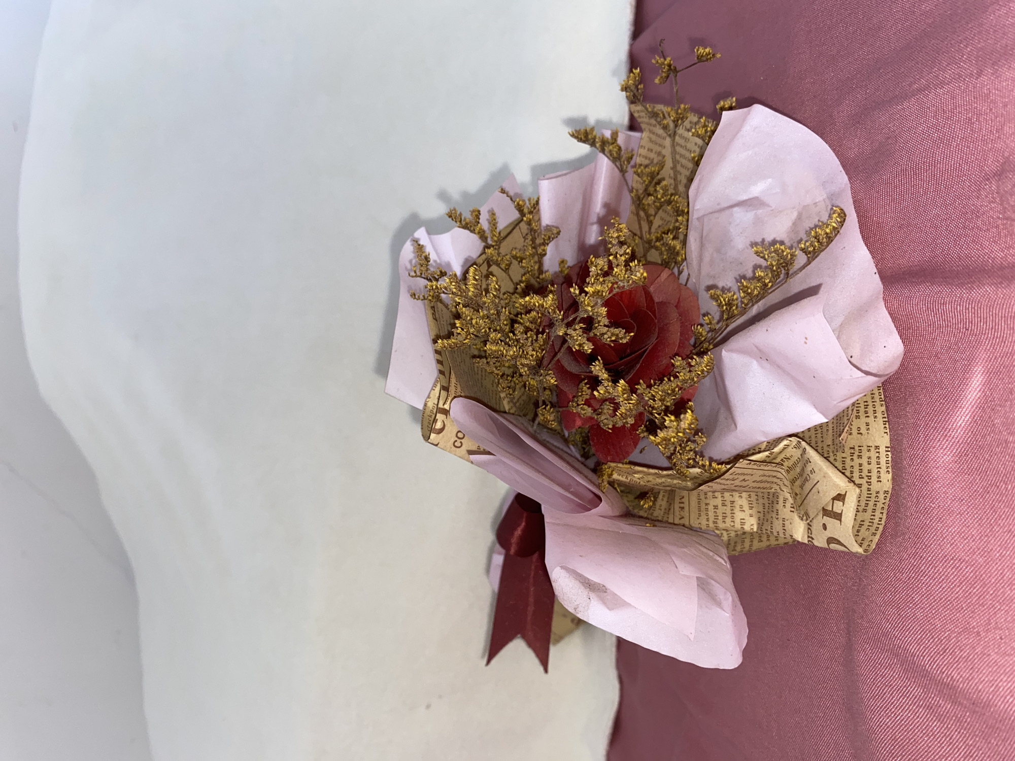 A bouquet of dried flowers, featuring a single red rose and small yellow blooms, is wrapped in pink tissue paper and newspaper. A burgundy ribbon is tied around the bouquet. The arrangement rests on a pink satin surface. The background is a softly lit, undulating white surface. The image is a close-up, studio-style product photograph, and no specific location in Davao City, Philippines, can be identified from the visual information provided.