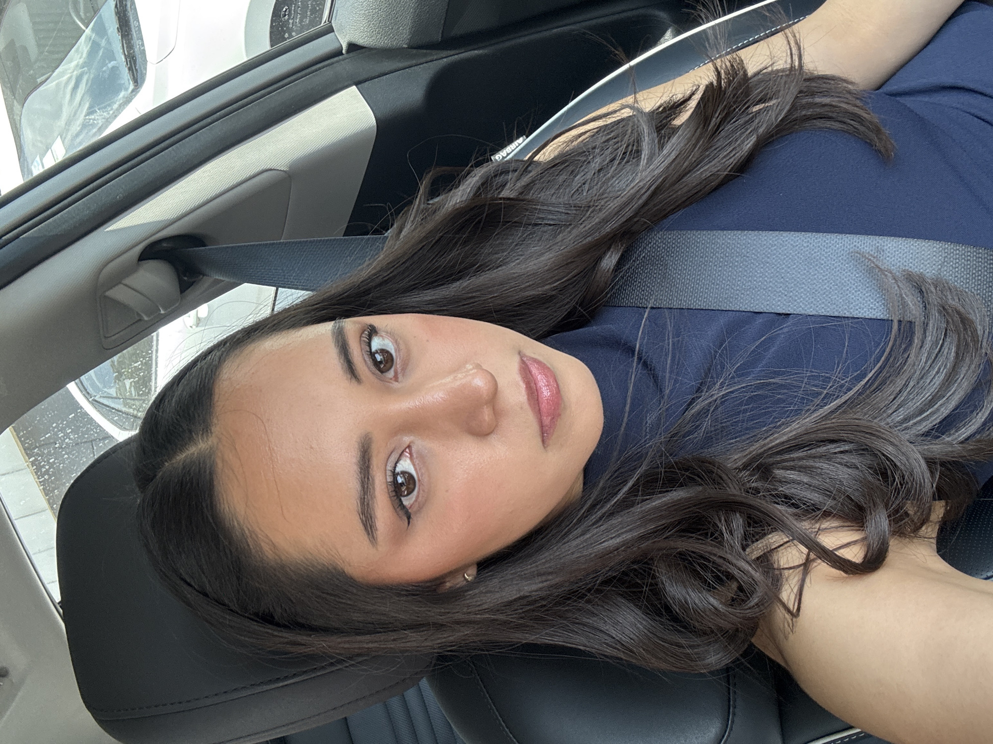 The image shows a woman inside a car, likely a sedan, wearing a dark blue shirt and a seatbelt. Her dark brown hair is styled with loose curls. She is looking directly at the camera. The car interior features a black leather seat. The location is purportedly Puebla, Mexico, but the image itself does not provide any explicit geographical markers to independently confirm this information. The car's interior and a partially visible exterior view of another vehicle through the window are the primary environmental elements captured.