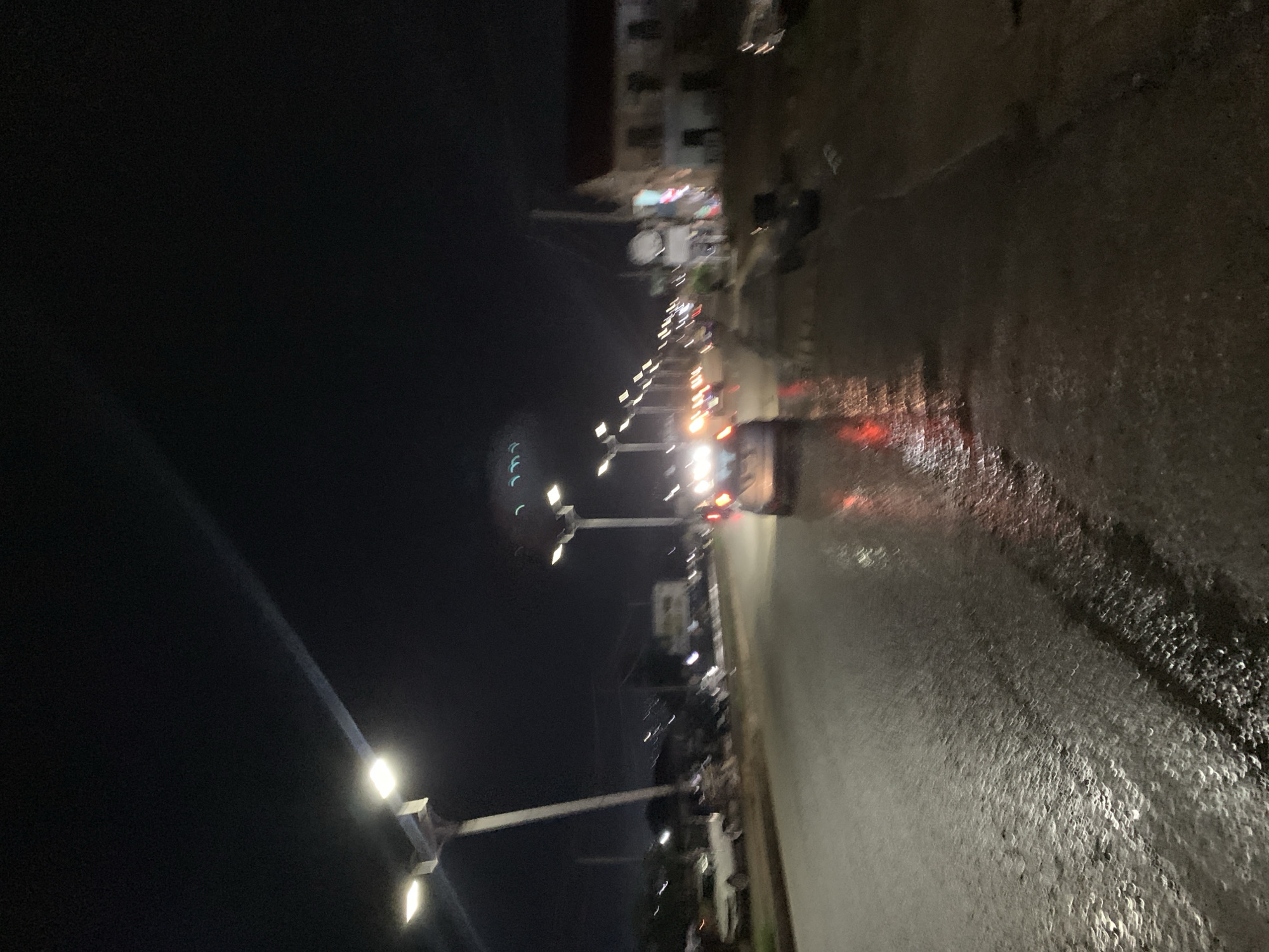 The image presents a night-time street scene in Rafin Par, Nigeria, viewed from the perspective of someone looking down a road. The sky is dark and clear, typical of night.
The most prominent feature is the wet, reflective asphalt road surface, which captures and elongates the intense lights from streetlights and vehicles. This wetness suggests recent rainfall or damp conditions. A row of modern streetlights with multiple bright fixtures lines the left side of the road, casting strong illumination and creating distinct reflections on the wet ground. These lights extend into the distance, guiding the eye along the street.
Vehicles are visible on the road, some appearing as streaks of bright light (red from taillights, white from headlights) due to motion blur or the long exposure of the photograph. One vehicle in the mid-ground shows discernible red taillights, while another, possibly a white car, is visible further down the street on the left.
Along the right side of the road, buildings line the street. These appear to be multi-story structures, some with visible windows and subtle interior lighting. There's a noticeable area with a lit commercial sign further down the street, though no text is legible. A large, rectangular white sign is also visible on the right side.
The overall impression is a vibrant yet somewhat blurry night landscape, characterized by the interplay of artificial lights and their reflections on the wet urban surface. No people are clearly visible, and there is no discernible text in the image.