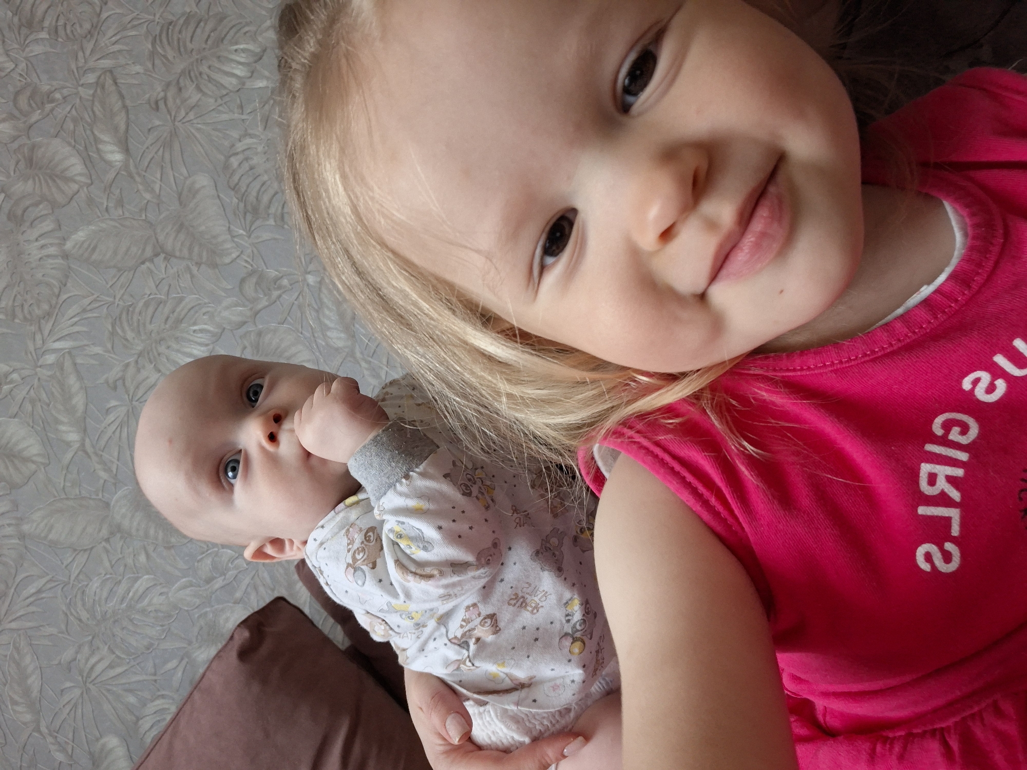 A baby and a young child are in the image. The baby is wearing patterned pajamas and is being held by someone. The young child has blonde hair and is wearing a pink shirt. They appear to be interacting or posing together. The background is a wallpapered surface.