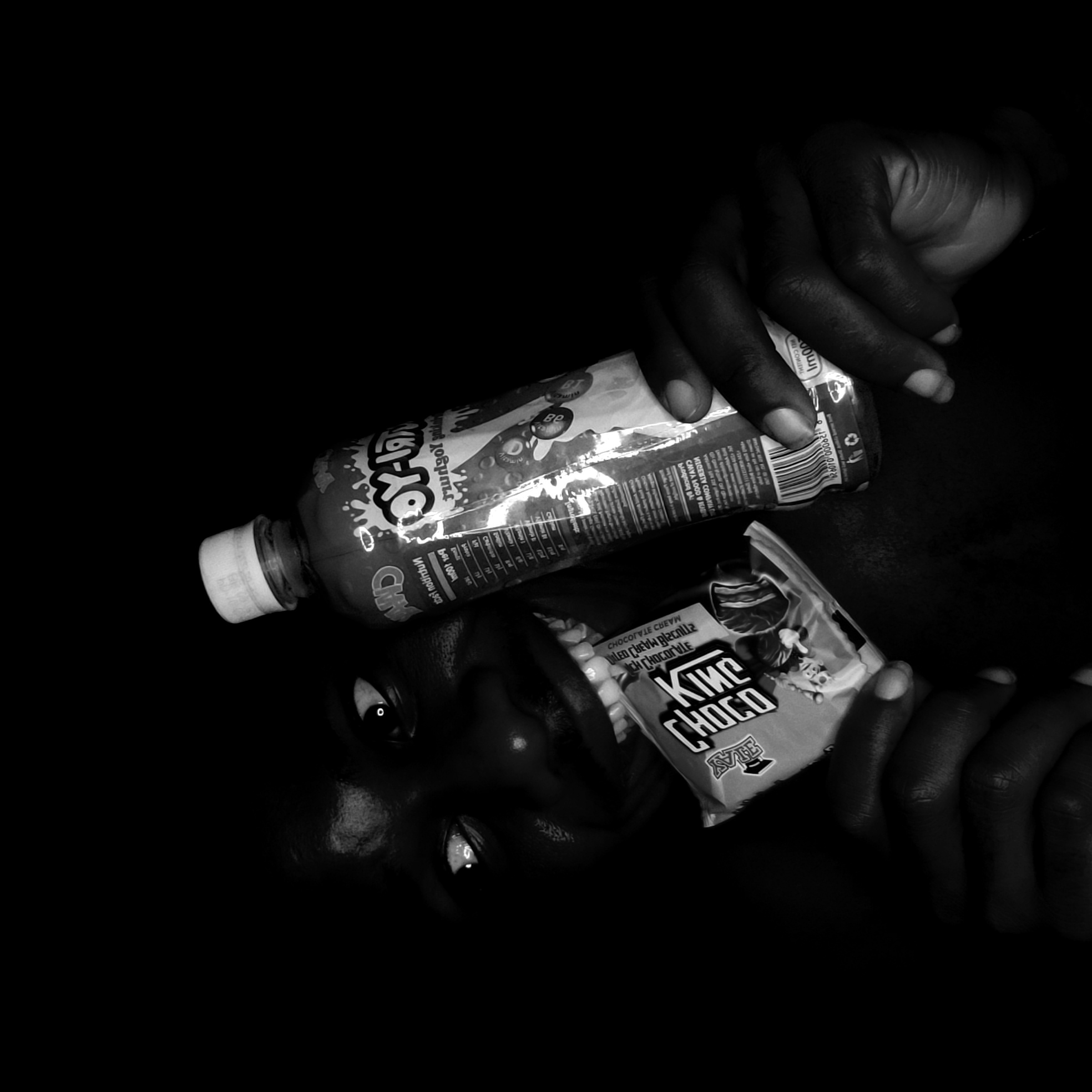 The image is a black and white photograph of a person lying on their back, holding a bottle of yogurt drink and a chocolate bar. The brand of yogurt drink is "Chudo Yogurt" and the chocolate bar is "King Choco". The person's face is visible, with eyes looking upwards. The hands are holding the bottle and chocolate bar close to the person's mouth. The background is black, creating a high-contrast image. Narayi, Nigeria is not mentioned in the image itself, nor is it possible to confirm that location based on the visual evidence.