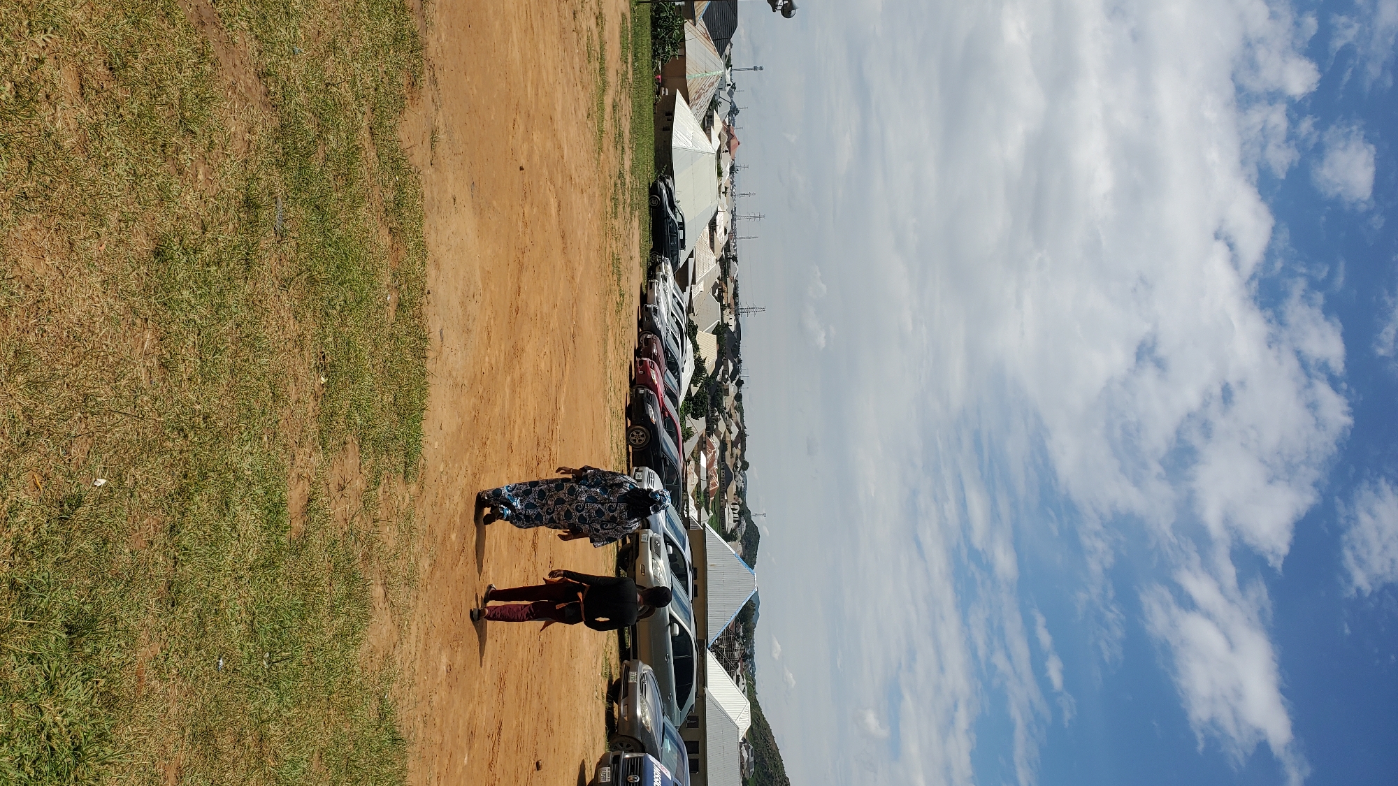 In Barakin Akawo, Nigeria, two people walk across a dry, dusty field towards a row of parked cars. The person in front wears a blue and white patterned garment, while the person behind them wears a black long-sleeved shirt and maroon pants. Beyond the cars, a cluster of buildings with distinctive conical roofs and white sheeting is visible, nestled against a backdrop of rolling hills under a partly cloudy sky. The scene suggests an outdoor gathering or event in a semi-urban area.
