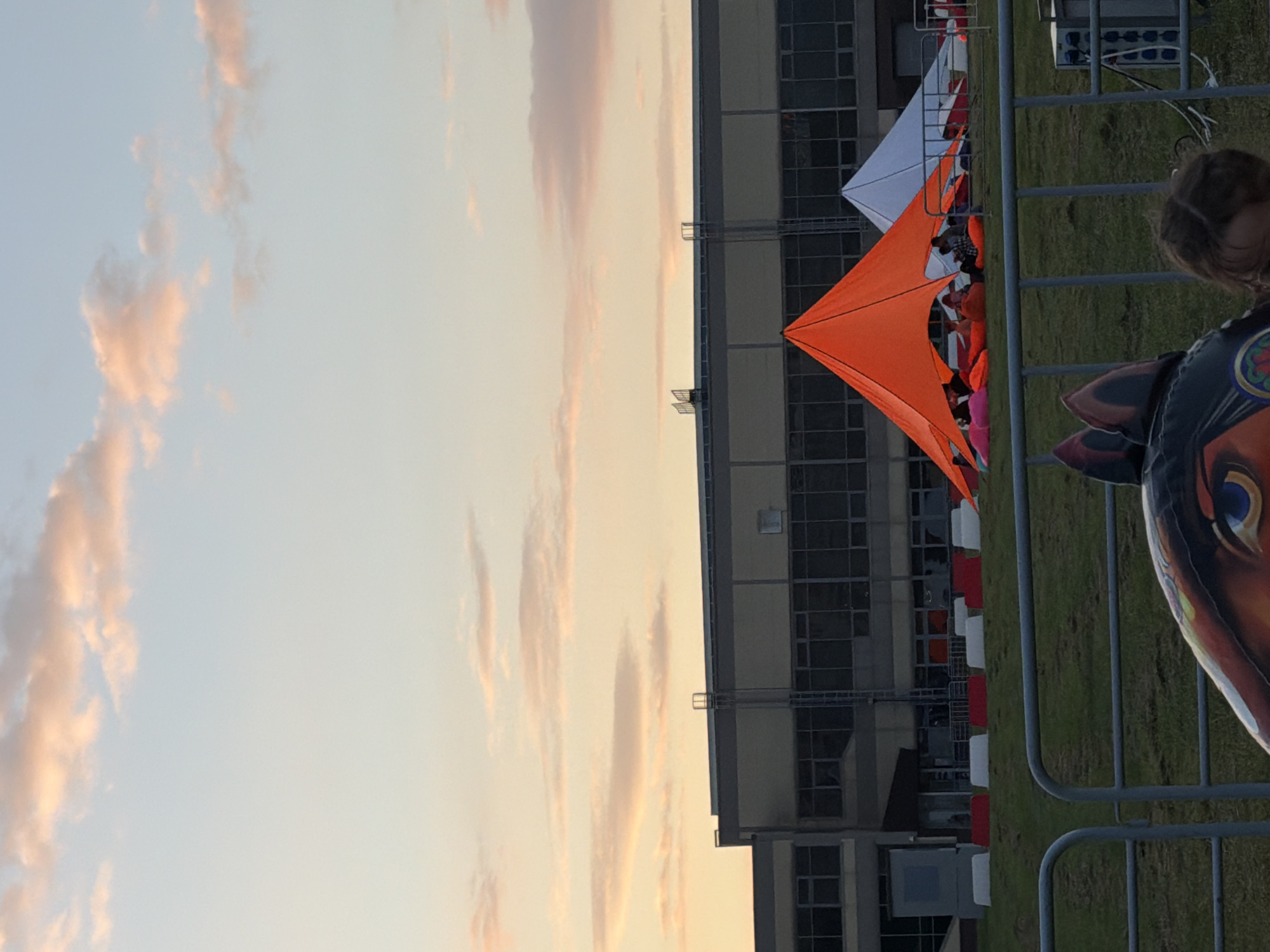 The image shows an outdoor scene at dusk. A long, low building with many windows is in the background. In front of the building, there are two tents, one orange and one white, with people sitting under them on cushions. A metal fence runs across the foreground, and behind it, a grassy slope leads up to the tents. In the very front of the image, a child's head is visible, looking down. The sky above is a pale blue with streaks of pink and orange clouds.