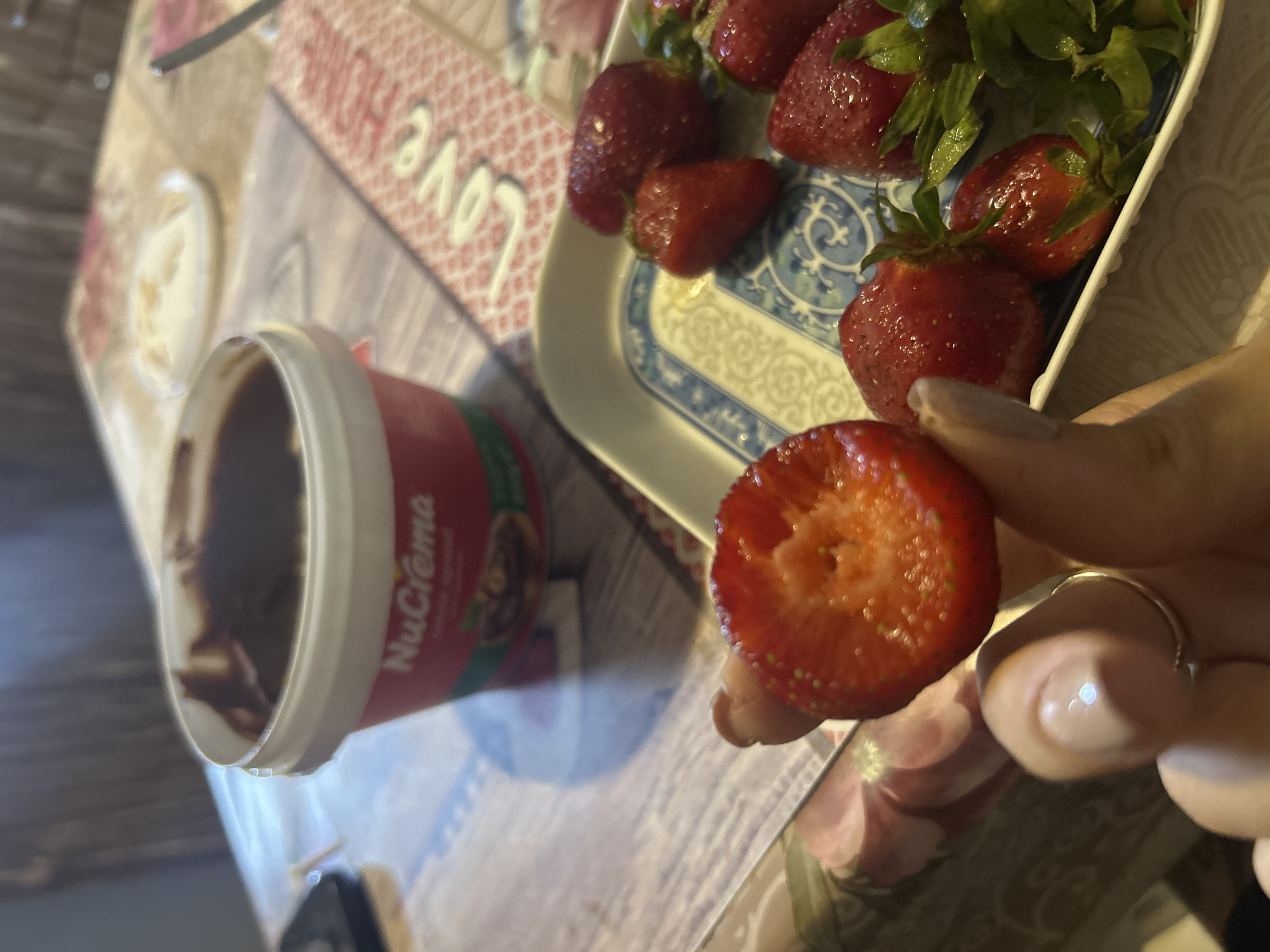Here is a detailed description of the image:
The image depicts a close-up, indoor scene of a person preparing to eat strawberries with chocolate hazelnut spread. A half-eaten strawberry is held in the person's fingers, exposing the inner texture. A tub of "NuCrema" chocolate hazelnut spread is visible to the left, suggesting it's intended as a dip. A platter with several fresh strawberries, some with green stems still attached, is positioned near the center. The background features a table surface decorated with a pattern that includes the word "love." The lighting is somewhat dim, casting shadows that add depth to the composition.