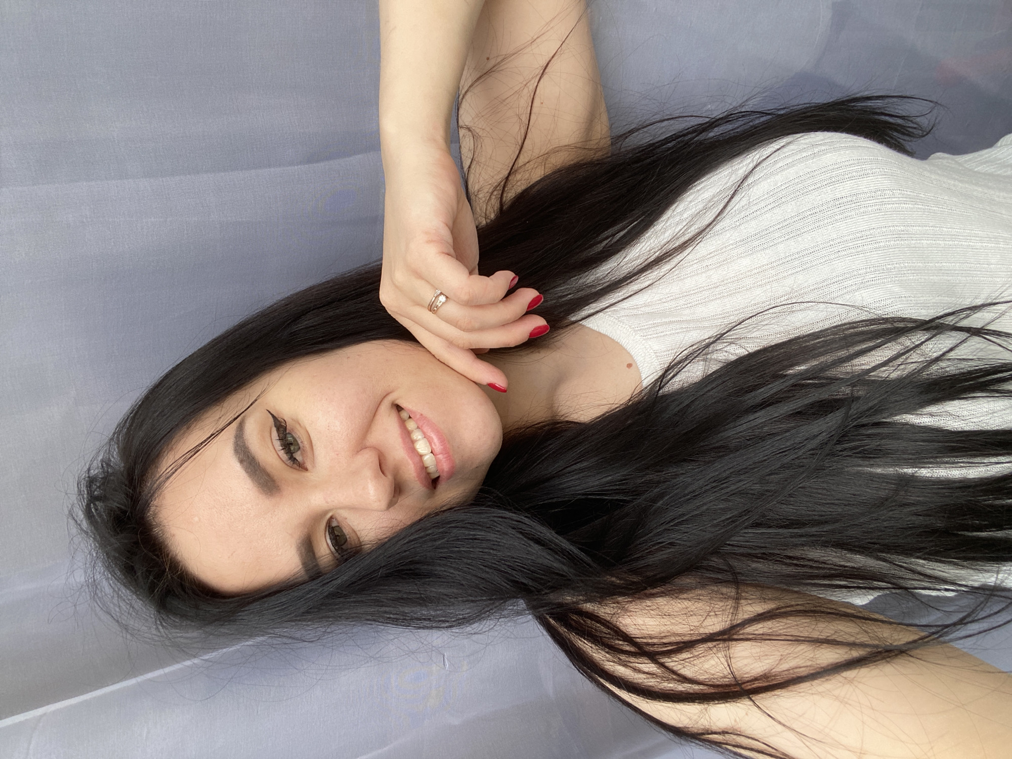 A young woman with long, straight, dark hair is depicted lying on her back, looking upwards towards the camera with a cheerful expression. Her head is angled slightly to her left, and she is smiling broadly, revealing her upper teeth. She has a fair complexion, dark eyebrows, and visible winged eyeliner. Her eyes are brown.
She is wearing a white, ribbed top. Her right arm is raised, with her hand positioned near her face, fingers gently touching her chin/cheek area. She wears red nail polish on her fingernails and two rings on the ring finger of her right hand: one a simple band and the other appearing to have a small stone. Her hair is fanned out around her head and shoulders, with strands extending across her arm and top.
The background consists of a light grey or possibly light blue textured fabric, likely a sheet or soft covering, suggesting an indoor setting such as a bed. The lighting appears soft and even, consistent with daylight indoors. There is no visible text in the image. The overall mood is relaxed and joyful. The location context is Chaikovsky, Russia.