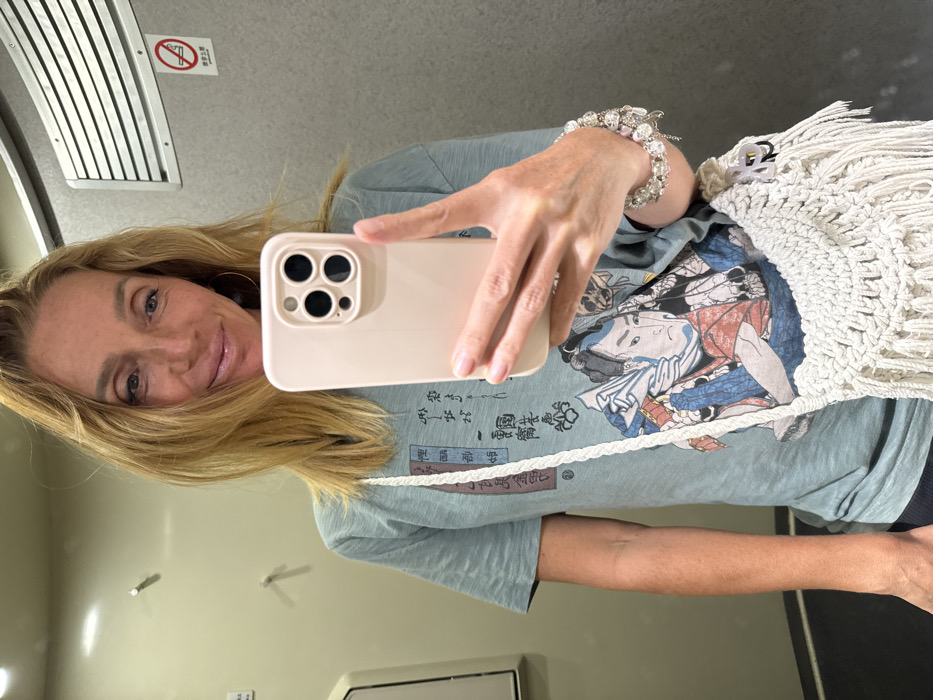 A mirror selfie captures a blonde-haired female subject in an indoor public space, identified as being in China by a visible "No Smoking" sign featuring Chinese characters (禁止吸烟). The specific city of Dong'an cannot be determined from the provided image.
The subject has light skin, blue eyes, and a slight smile, looking directly at the camera. Her long, blonde hair is parted to her left. She wears a short-sleeved, light blue-green t-shirt adorned with a multi-colored ukiyo-e style print depicting a male figure and vertical Japanese text. A white, braided-strap macrame bag with fringe details is slung over her left shoulder. A multi-beaded bracelet is visible on her left wrist.
Her right hand holds a light pink or beige smartphone, equipped with a triple-lens camera array, positioned in front of her mid-torso, capturing the reflection.
The background features light-colored wall panels. A metallic horizontal vent is visible in the upper right, positioned above the "No Smoking" sign. A white, wall-mounted fixture is partially visible on the left side of the frame. The environment is uniformly lit by artificial indoor lighting.