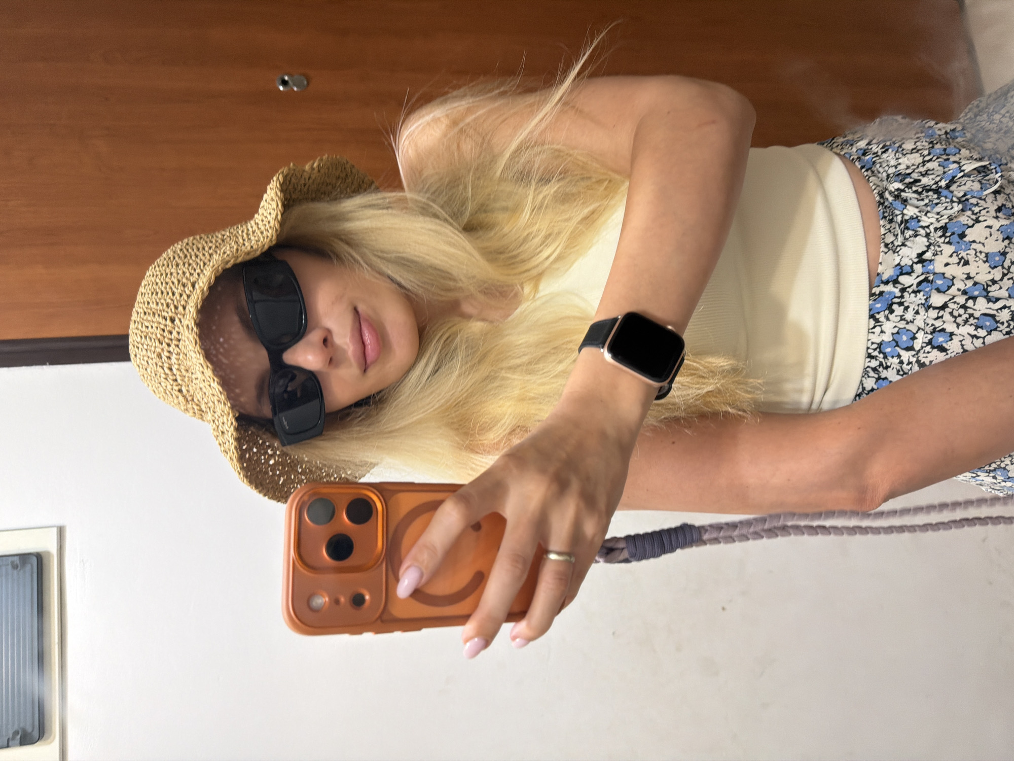 In Budva, Montenegro, a young woman with long blonde hair, wearing a straw hat, sunglasses, a light-colored ribbed tank top, and floral-patterned shorts, is taking a mirror selfie. She is holding an orange smartphone in her left hand, which is adorned with a silver ring on her finger. A dark smartwatch is visible on her right wrist. The background consists of a plain white wall on the left and a wooden door with a silver doorknob on the right. The scene appears to be indoors, possibly in a residential setting or hotel room, given the simple wall and door.