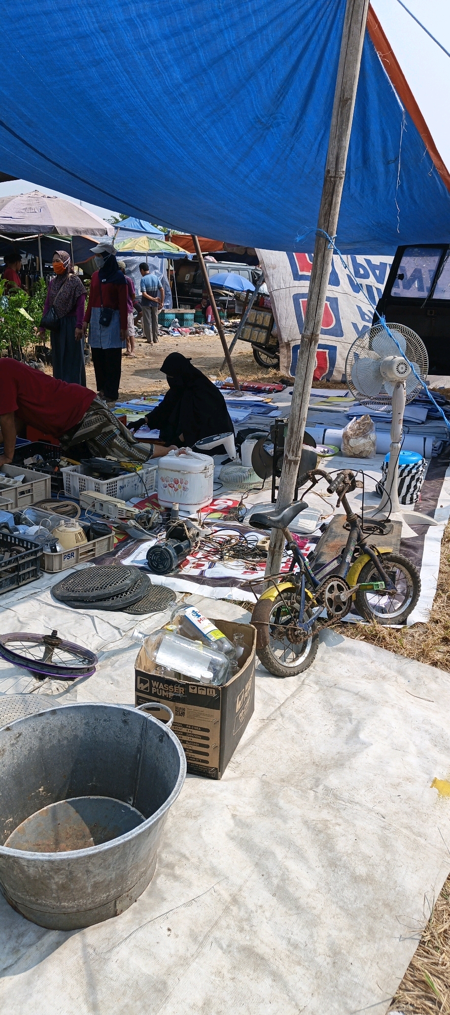 The image depicts an outdoor market scene under a large blue tarp, likely in Jatinom, Indonesia. Several people are visible in the background, some wearing headscarves and masks. The foreground is covered by a white sheet upon which various items are displayed for sale. These include what appears to be electronic components, a rice cooker, a fan, a bicycle, and a large metal bucket. A person dressed in black is seated on the sheet, attending to the items. The overall impression is of a busy, informal marketplace.