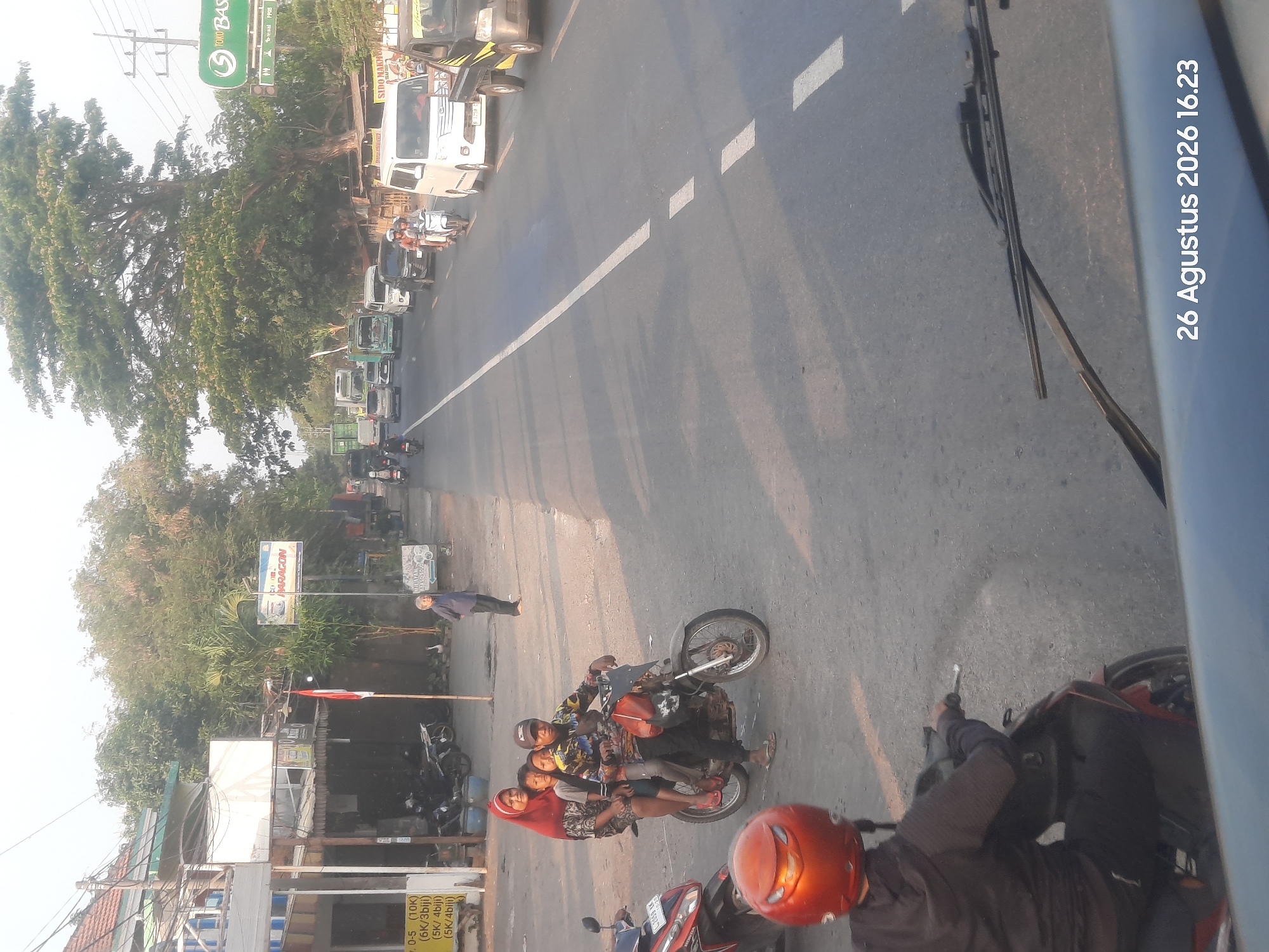 The image depicts a street scene in Sukokerto, Indonesia, on August 26, 2026, at 16:23. The perspective is from inside a vehicle, looking out through the windshield and side window. The road is asphalt and marked with white dashed lines. Sunlight casts long shadows from trees onto the road, indicating late afternoon.
In the foreground, a person wearing a black hooded jacket and an orange helmet is riding a red motorcycle. To their left, another red motorcycle is parked. Further ahead on the right side of the road, a group of four people, including a woman wearing a red headscarf and two children, are riding on a single motorcycle. The driver, a male wearing a cap, is at the front.
On the left side of the road, there are buildings with shops and signage, including what appears to be a price list. A few motorcycles are parked in front of these establishments. An Indonesian flag is prominently displayed.
The road is moderately busy with traffic. Vehicles visible include several cars, white vans, and trucks. A black pickup truck is in the right lane. In the distance, more vehicles are seen moving along the road, with trees and vegetation lining the sides. A large, mature tree with dense foliage dominates the upper right portion of the image. Several utility poles are also present. A green sign with white text, likely for a business, is visible on the right.