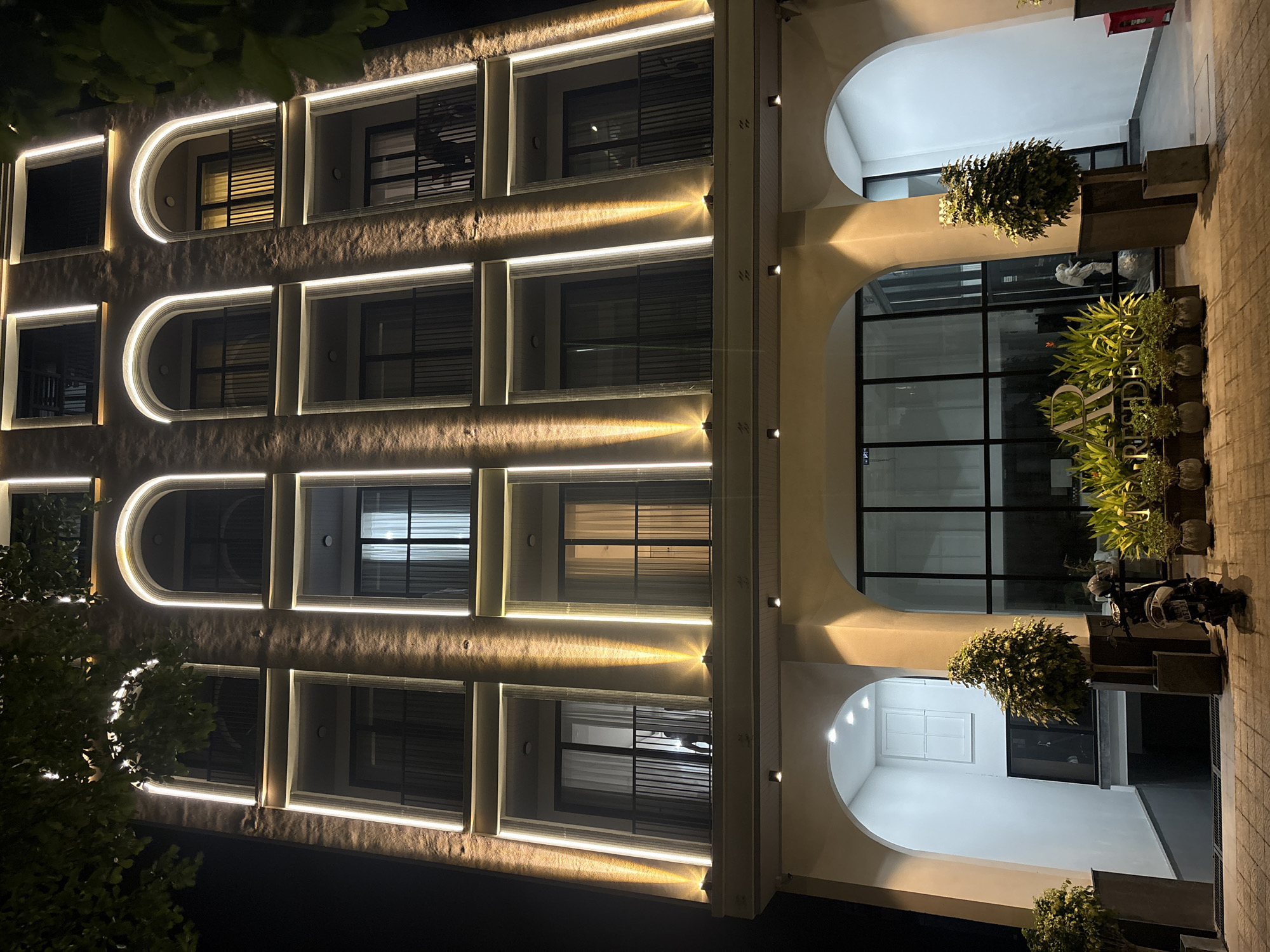 The image depicts the exterior of a modern building at night in Đà Nẵng, Vietnam. The building features a textured facade with rows of windows, each framed by curved, illuminated accents. The lighting creates a dramatic and inviting ambiance.
The scene is primarily focused on the architecture, with prominent arched entrances on the ground floor. These entrances lead to interior spaces with large windows and a modern aesthetic. Several potted plants and trees are strategically placed around the building, adding a touch of greenery to the urban setting.
Visible elements include a motorcycle parked near one of the entrances, suggesting a commercial or residential establishment. There are also indications of people, though they are not clearly defined, giving a sense of activity without explicit focus.
The time of day is clearly night, as indicated by the dark sky and the artificial lighting illuminating the building's features. The weather appears clear, as there are no visible signs of rain or overcast conditions. The overall mood is sophisticated and contemporary, highlighting the building's design and its nighttime illumination.
While the image is primarily architectural, a sign with partial text is visible on the facade. The visible letters appear to be "RESID" and "CHIC", hinting at a name or branding related to accommodation or a stylish establishment.