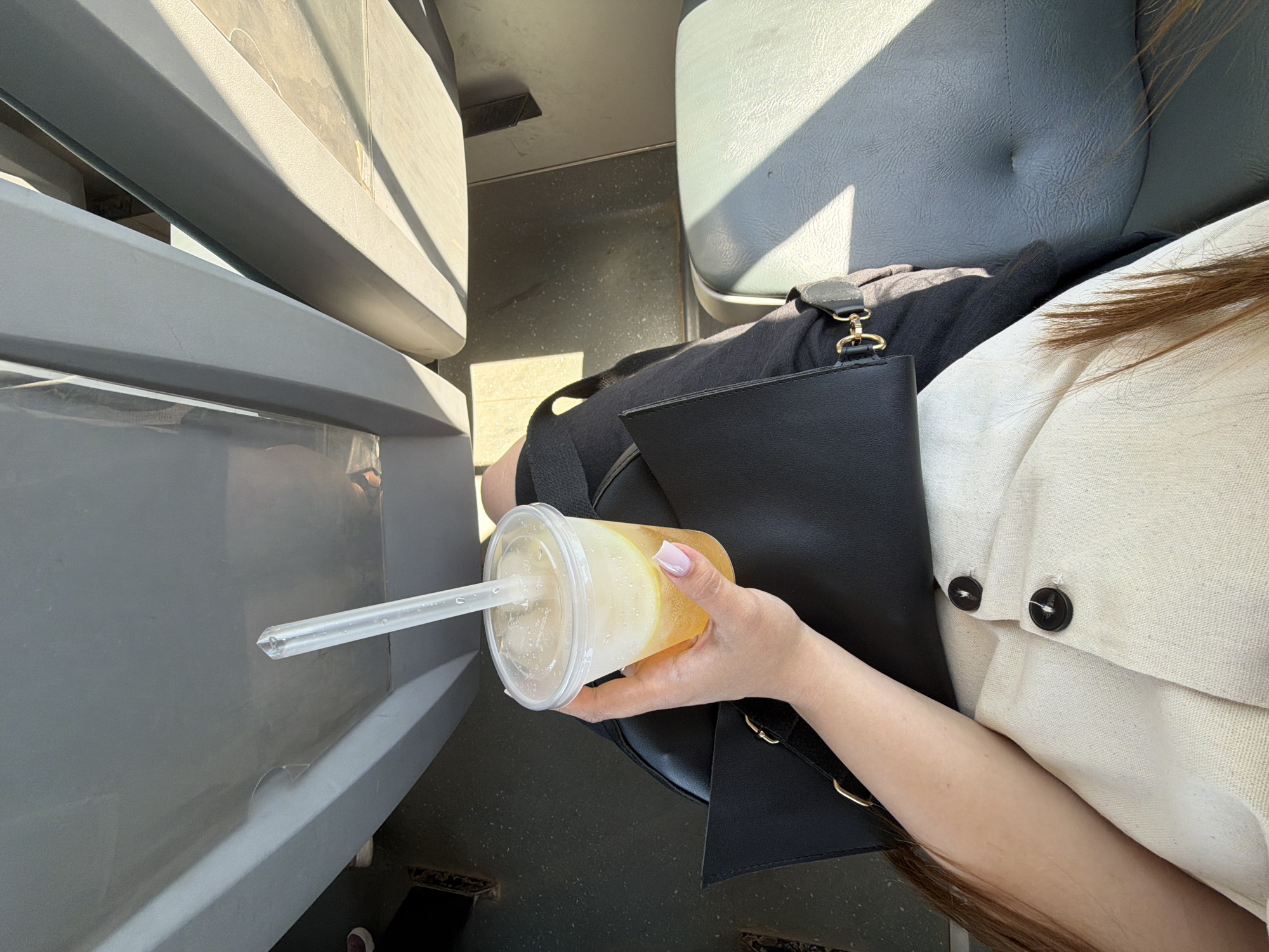 A high-angle shot shows a person holding a plastic cup filled with a light-colored beverage with a clear straw, resting on a black purse and black clothing. The person wears a white button-down shirt with two visible black buttons. The location appears to be indoors, possibly a bus or train, given the presence of grey seating and what looks like the side of another seat. The exact location in Chaikovsky, Russia, is impossible to determine from the given image, as it lacks contextual landmarks or location indicators. The focus is on the beverage and the person holding it.