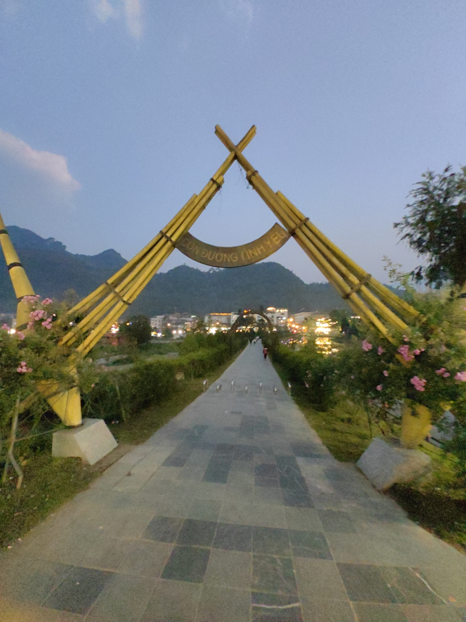 An eye-level, medium-shot photograph depicts a paved pathway leading through a large, arch-like structure at dusk in Sa Pa, Vietnam. The prominent archway, constructed from yellow, bamboo-like elements arranged in a stylized, pointed roof design, spans the pathway in the foreground. A curved sign bearing the Vietnamese text "CON ĐƯỜNG TÌNH YÊU" (The Love Road/Path) is affixed to the arch's upper section. Pink flowers and green foliage adorn the base of the archway on both sides.
The pathway, composed of alternating light and dark grey rectangular paving stones, extends straight into the mid-ground. A lone individual, dressed in red, walks away from the viewer down the distant portion of the path. To the right of the path, a body of water reflects numerous bright lights from buildings on its far bank. Beyond the water, a cluster of illuminated structures indicates a populated area, likely part of Sa Pa town. Dark, undulating mountains are visible on the horizon, silhouetted against a deep blue twilight sky with faint cloud formations. The overall scene suggests a pedestrian area within a mountainous, developed region.