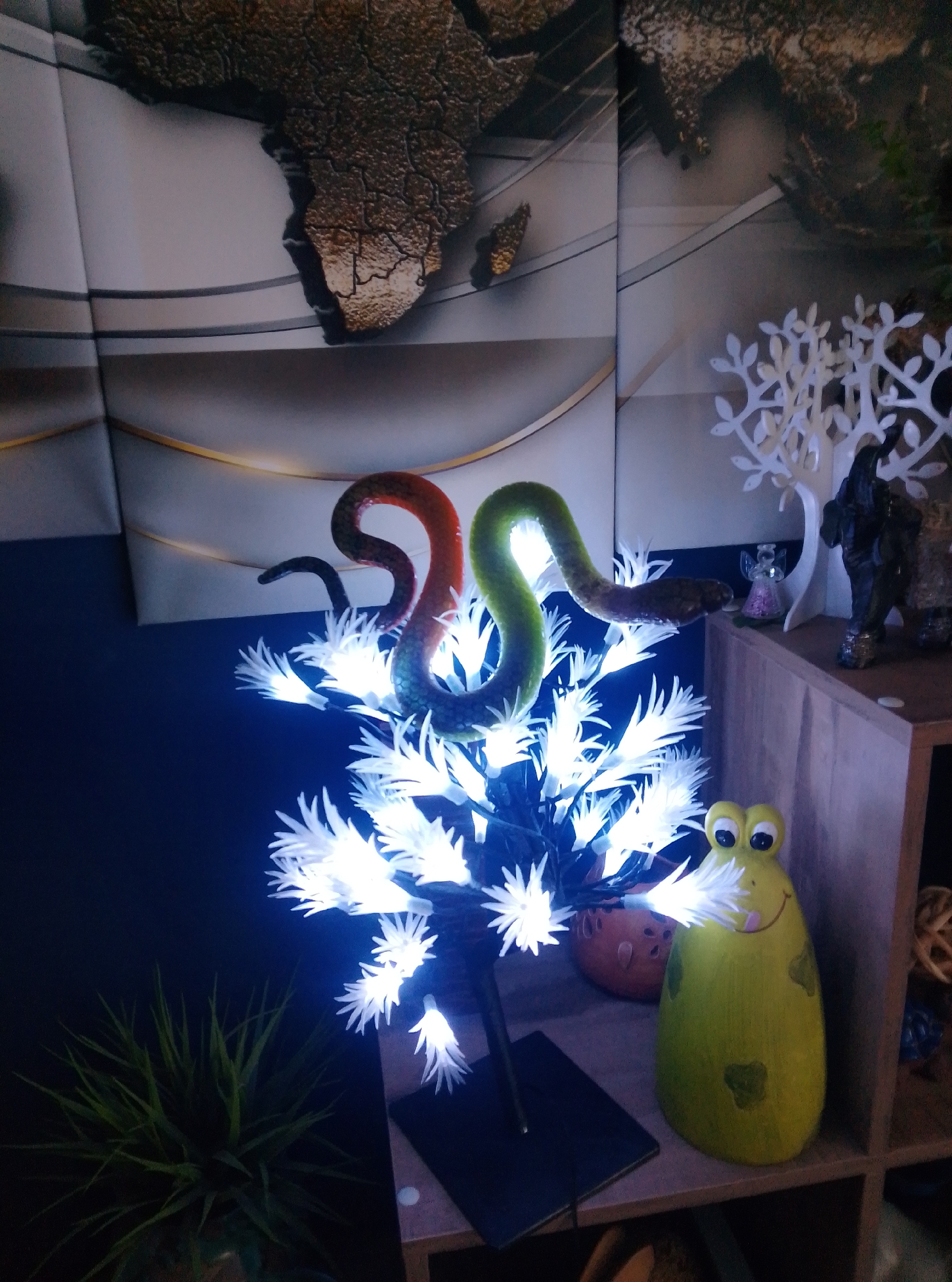The image depicts an indoor scene, likely a living room or entertainment area, decorated with a collection of items. Dominating the foreground is a decorative tree-like light fixture with numerous bright white LED lights, resembling blossoms or leaves. Coiled around the branches of this light are two artificial snakes, one orange and red, the other green. To the right of the light fixture, on a wooden shelf, sits a large, lime-green ceramic figurine of a frog with a smiling face and large eyes. Behind the frog and the light fixture, a large wall art or mural featuring a stylized map of Africa is visible. Further to the right on the shelf are additional decorative items, including a small white tree sculpture and a figurine of an elephant. In the lower left corner, a potted plant with spiky green leaves adds a touch of greenery to the composition. The lighting suggests an evening or dimly lit environment, with the LED tree being a primary light source. The overall impression is one of eclectic and whimsical decoration. This scene is located in the city of Wedel, Germany.